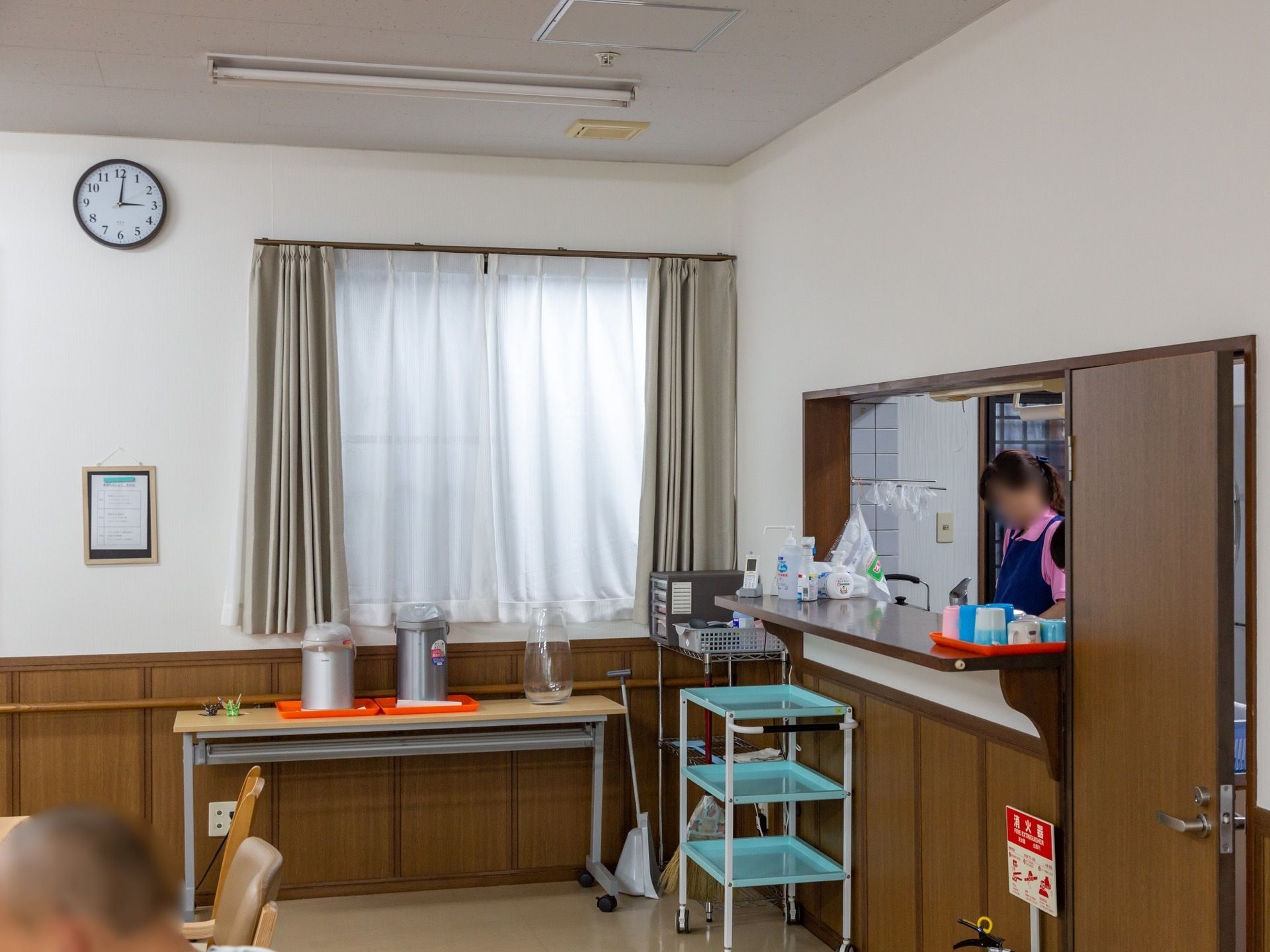 ブラウンの落ち着いた色合いのテーブルが並び、キッチンではスタッフが食事の準備をしている。窓の近くにはお湯の入ったポットがある。