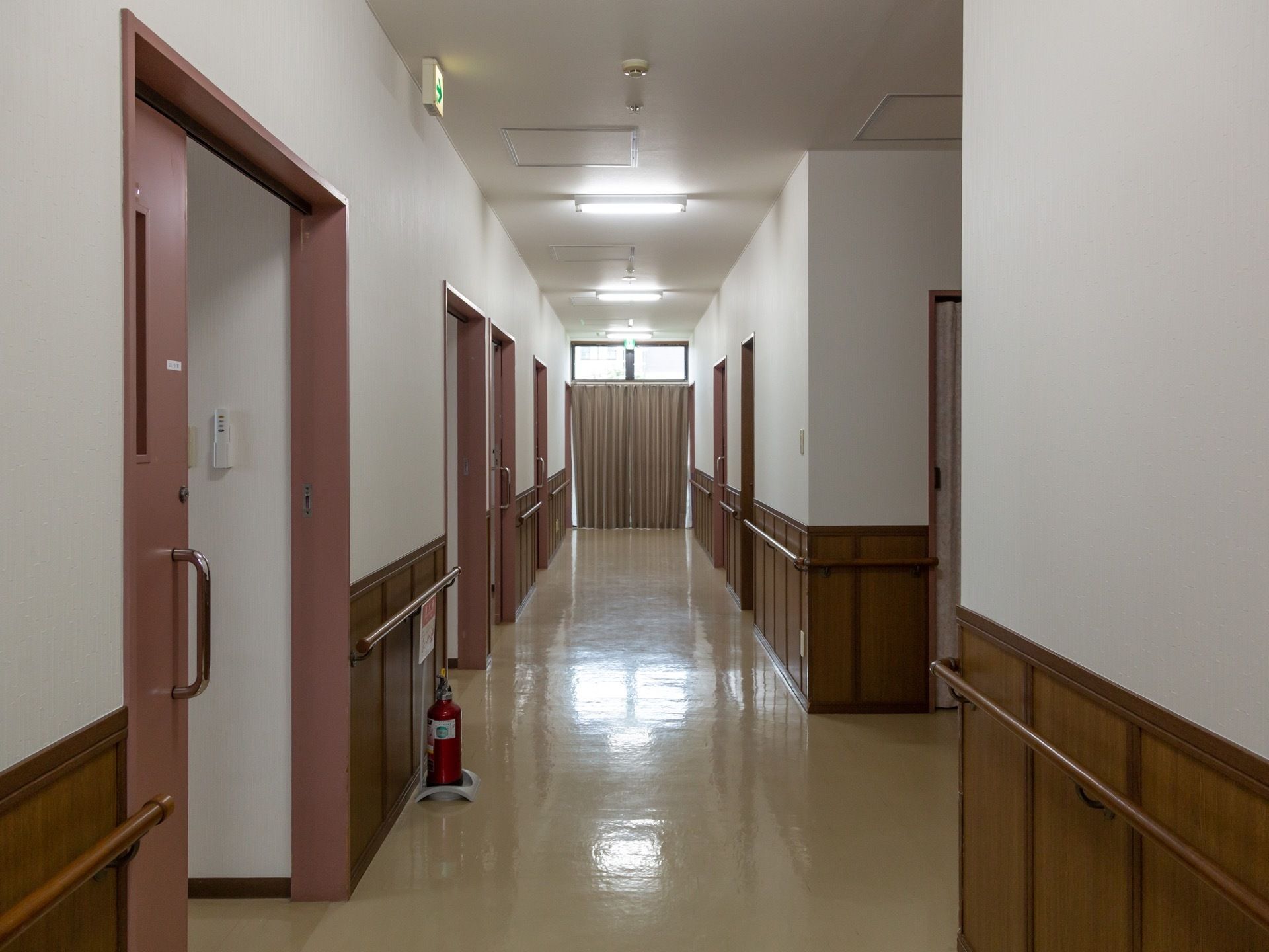 施設内の廊下には消火器が設置されている。天井近くには非常口の標識もある。居室に通じるドアはスライドタイプである。