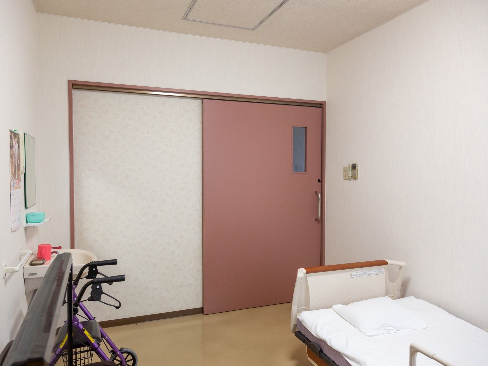 居室の扉は落ち着いたピンク色である。縦型の明かり窓もある。洗面台やベッド、歩行器などが備えつけられている。