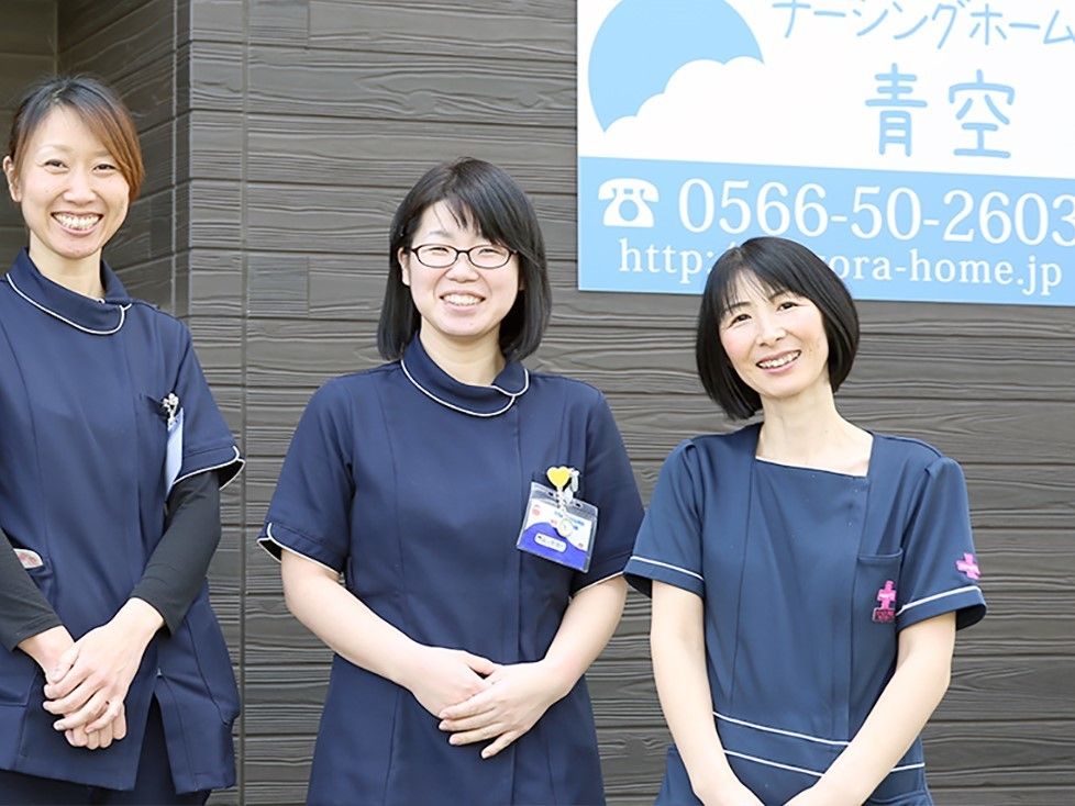 黒い制服を着た女性スタッフが３人並んで笑顔で写っている。ダークブラウンの外壁に施設名が書かれた表札が取り付けられている。