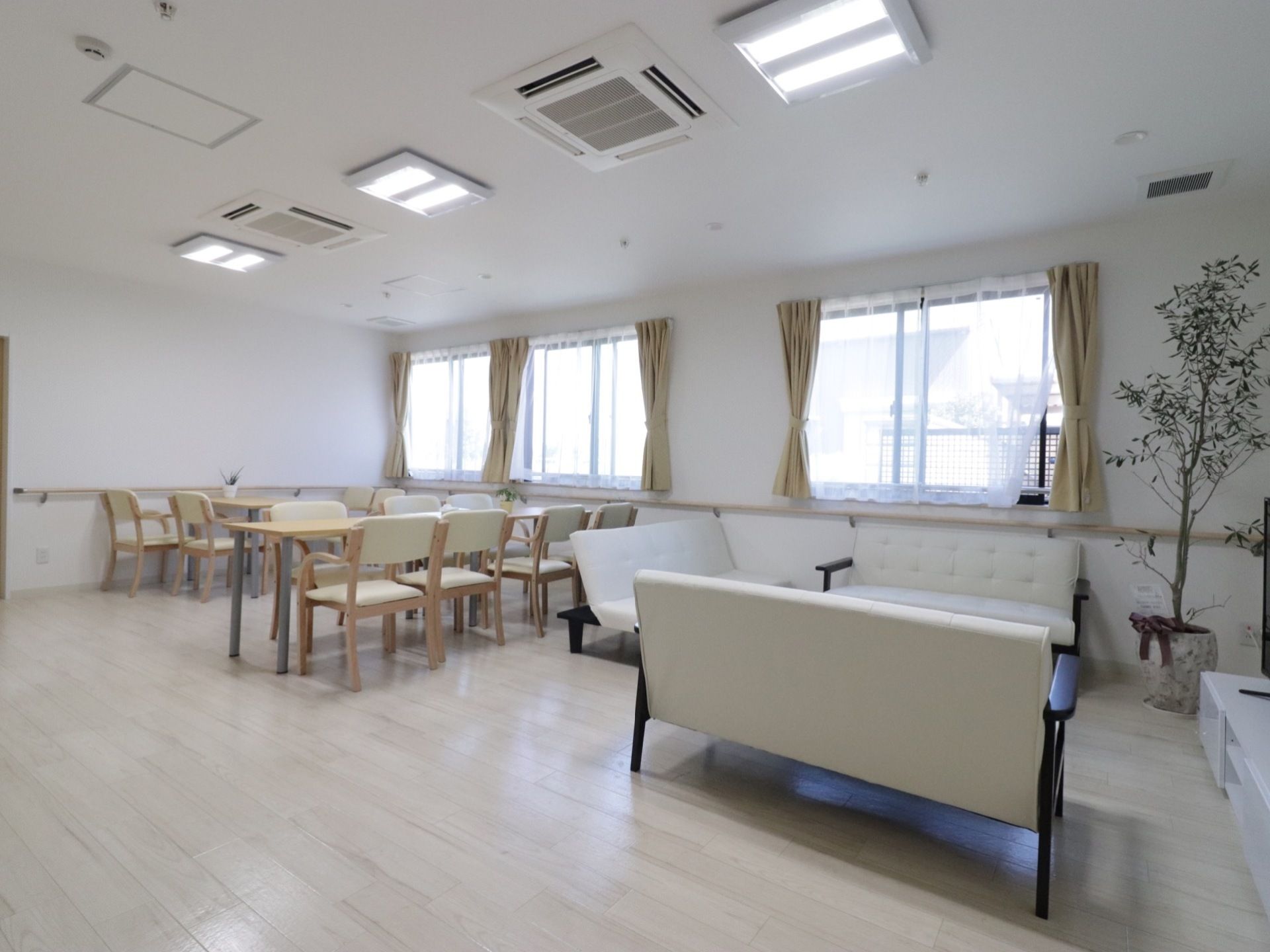 明るく開放的なリビングである。４人掛けテーブルやソファ、観葉植物が置かれている。壁に沿って手すりが取り付けられている。