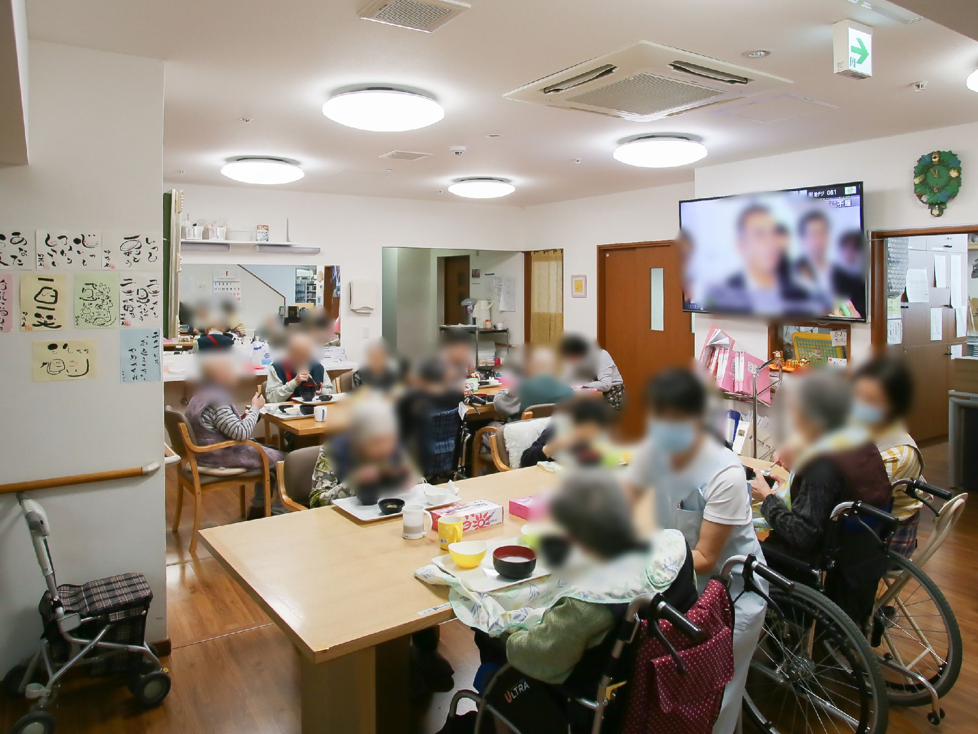 大きなテーブルが２つあり、入居者様が食事をしている。手前にいる車椅子に座った入居者様を横でスタッフがサポートしている。