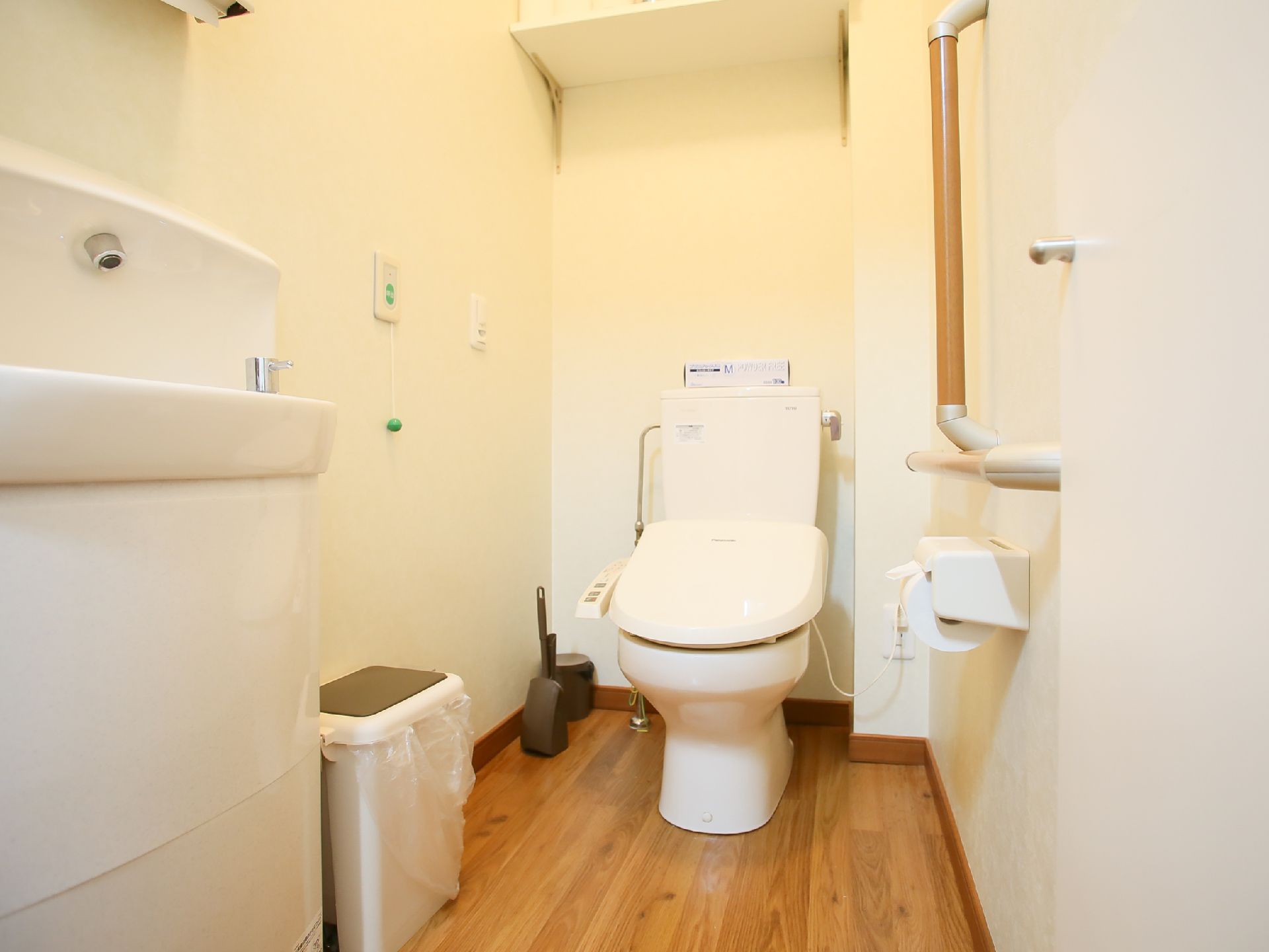 トイレの床はフローリングで手前に手洗い器が設置されている。壁にグリーンの緊急コールが取りつけられている。