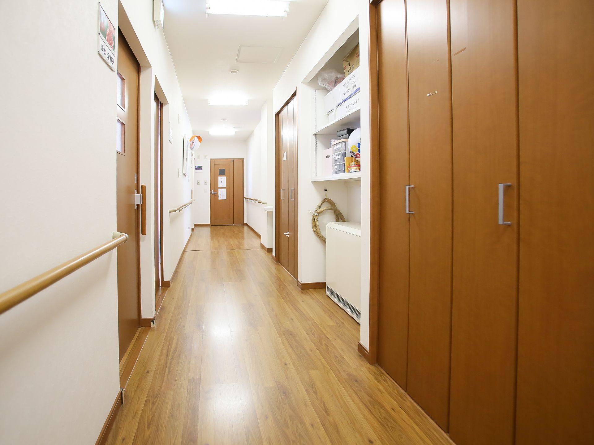 通路の片側に木目調の収納扉があり、その横には白い壁面収納がある。突き当たりにカーブミラーが設置されている。