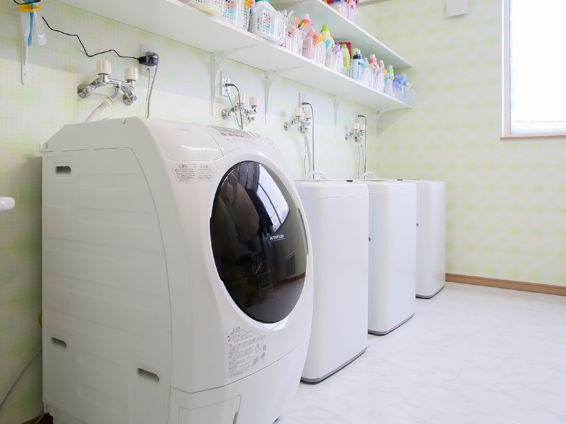 壁際に洗濯機が４台並んでいる。その上には２段の白い棚があり、洗剤などの入った白いバスケットが並んでいる。