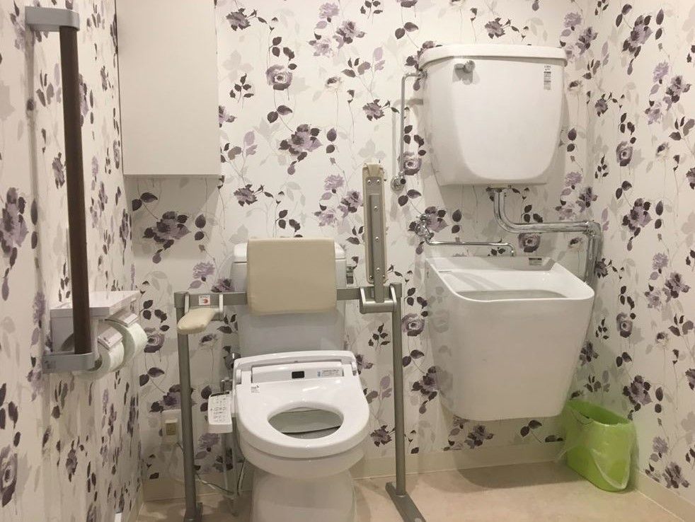背もたれや肘置き、オストメイト対応のトイレを設置した個室がある。グレーの濃淡で表現した柄の壁紙が貼られている。