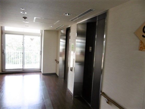 エレベーターの淵は銀色である。ホールの床はダークブラウンの板がはってある。近くに窓がついており、自然光が差し込む。
