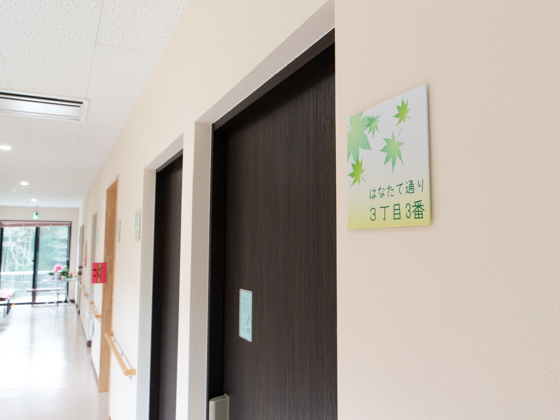 手すりが備わっている廊下である。ドアはこげ茶色の大きな横開きの扉になっている。その横の壁には各居室名が書かれた札がつけられている。