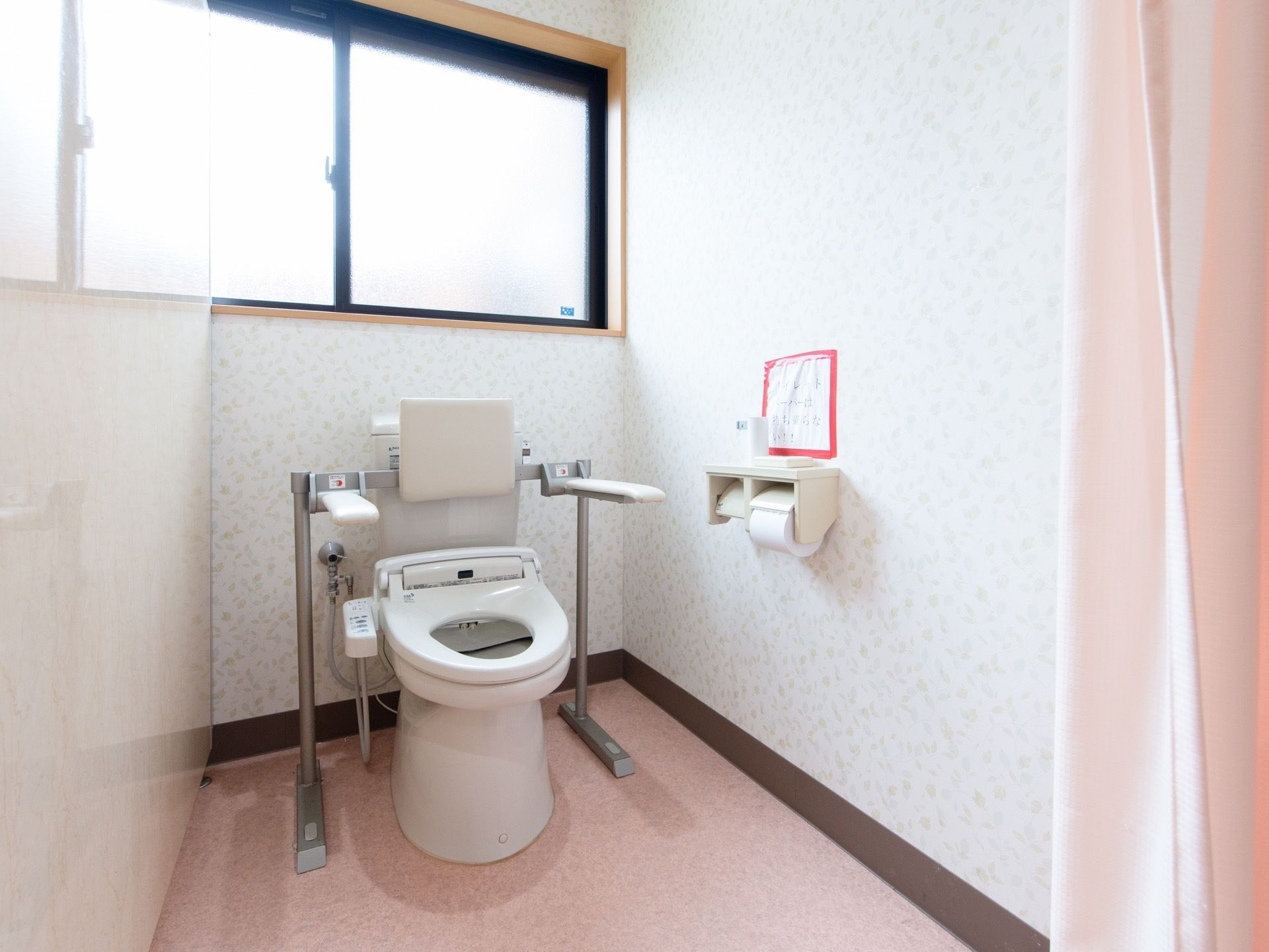 大きなすりガラスの窓があるスペースにトイレがある。背もたれや、便座の左右に可動式の白い大きな手すりがついている。