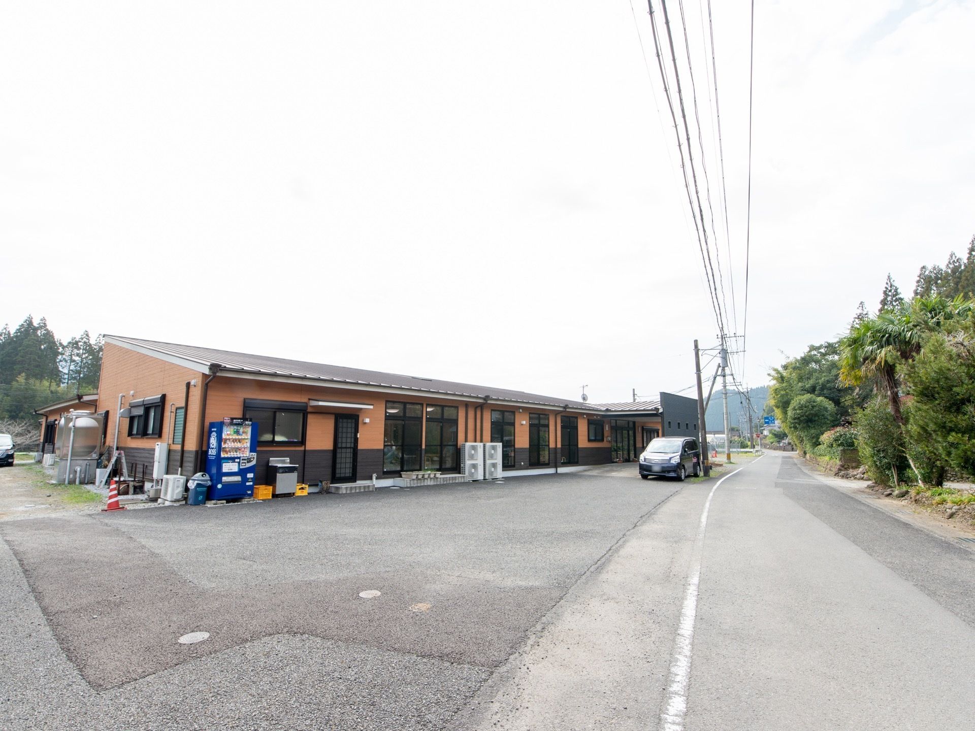 すぐ横を道路が走っている。窓がたくさん備わっている。屋外には自動販売機が置かれている。自然に囲まれている。