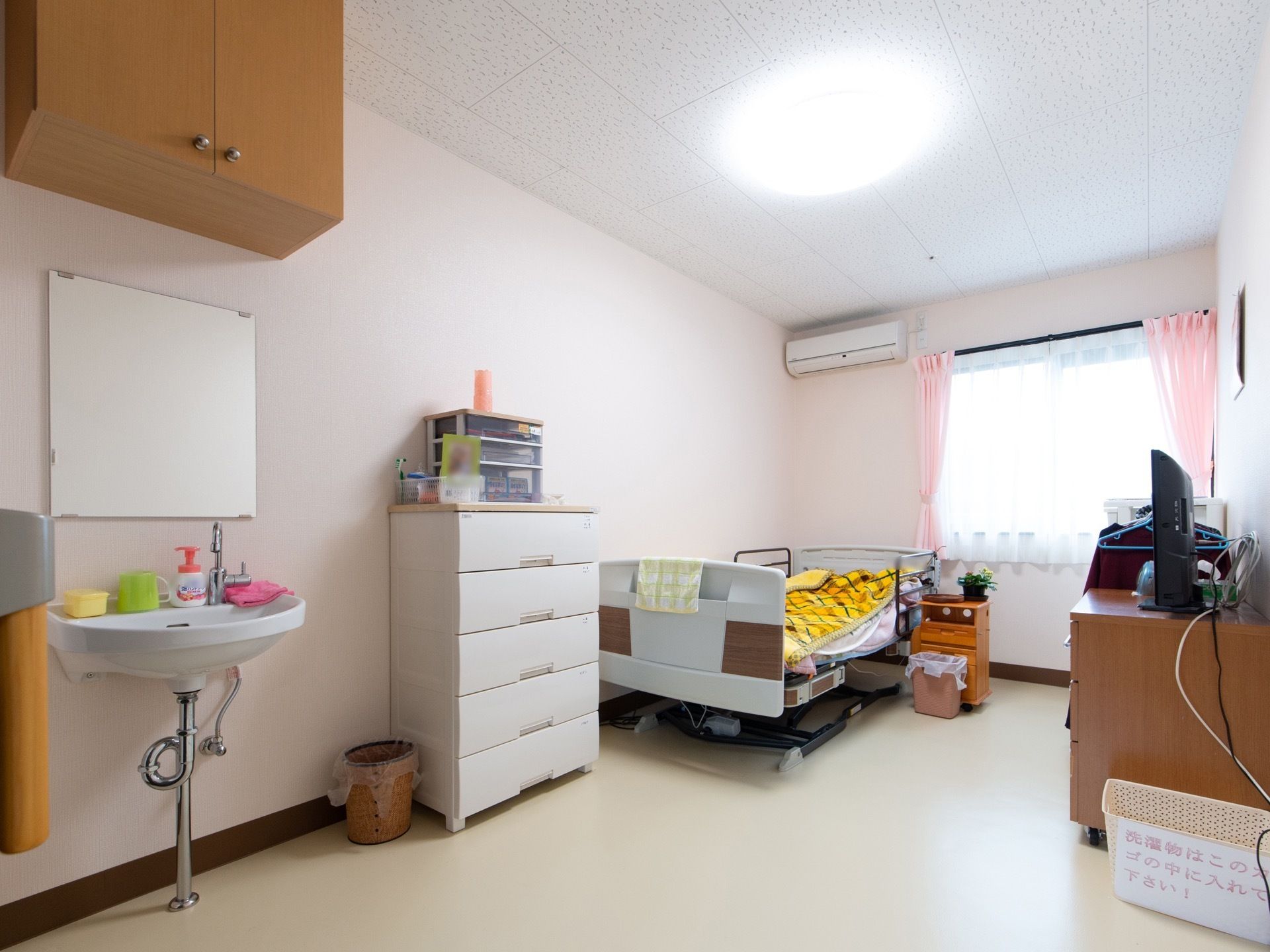 大きな鏡のついた洗面台が設置されている。窓が大きく明るく開放的である。ベッドやタンス、テレビが置かれている。