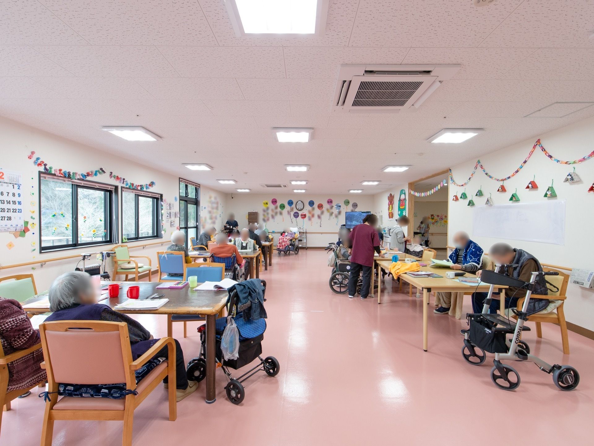 壁にはカレンダーや創作品が飾られている。たくさんのテーブルや椅子が置かれている。たくさんの入居者様がくつろいでいる。