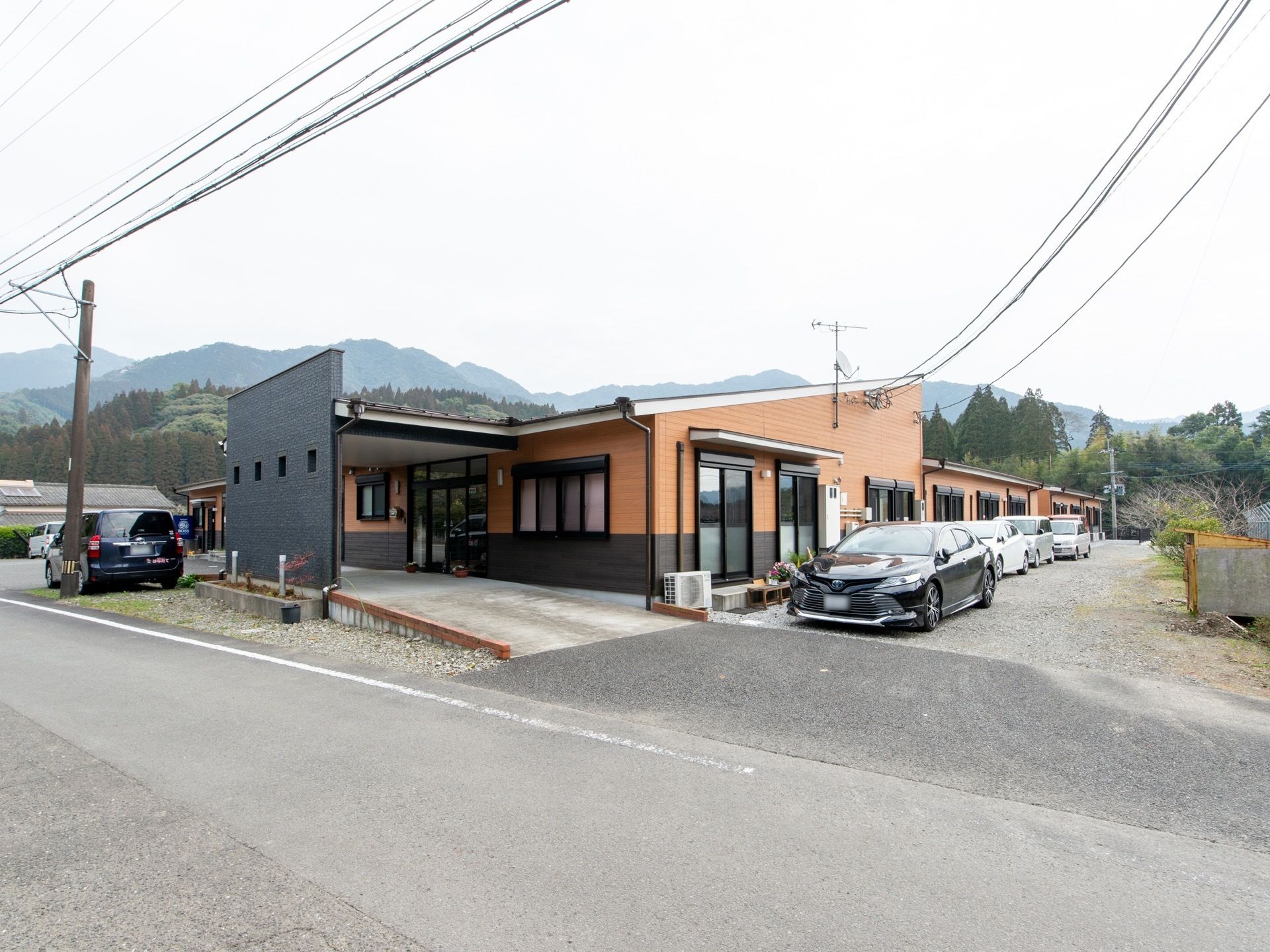 平屋建ての建物で、前方には道路が通っている。建物の横は駐車スペースとなっており、車が複数停まっている。