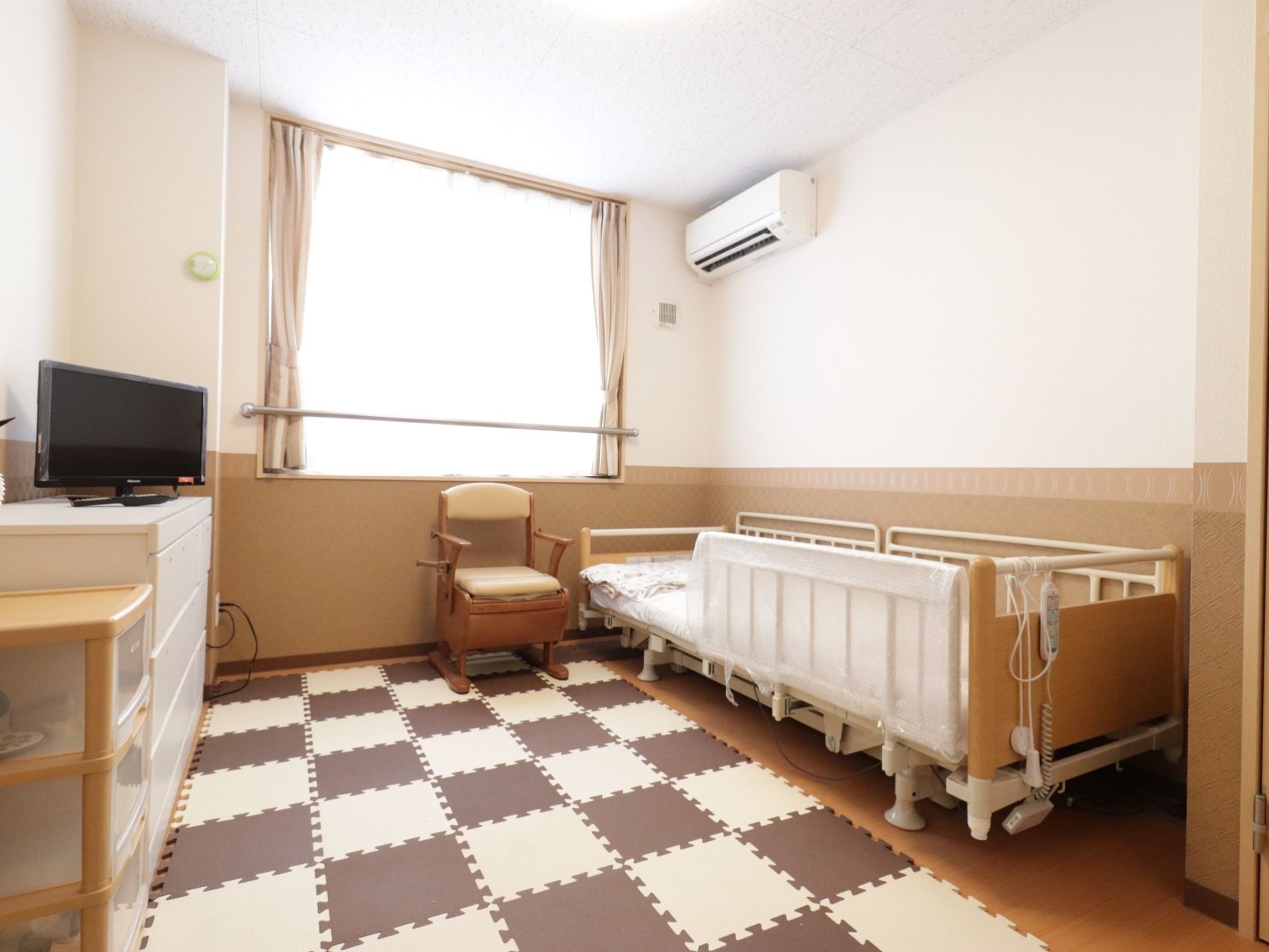 一人用居室の風景。ベッドサイドには、家具調のポータブルトイレや衣装ケースやテレビが置かれている。エアコンも設置されている。