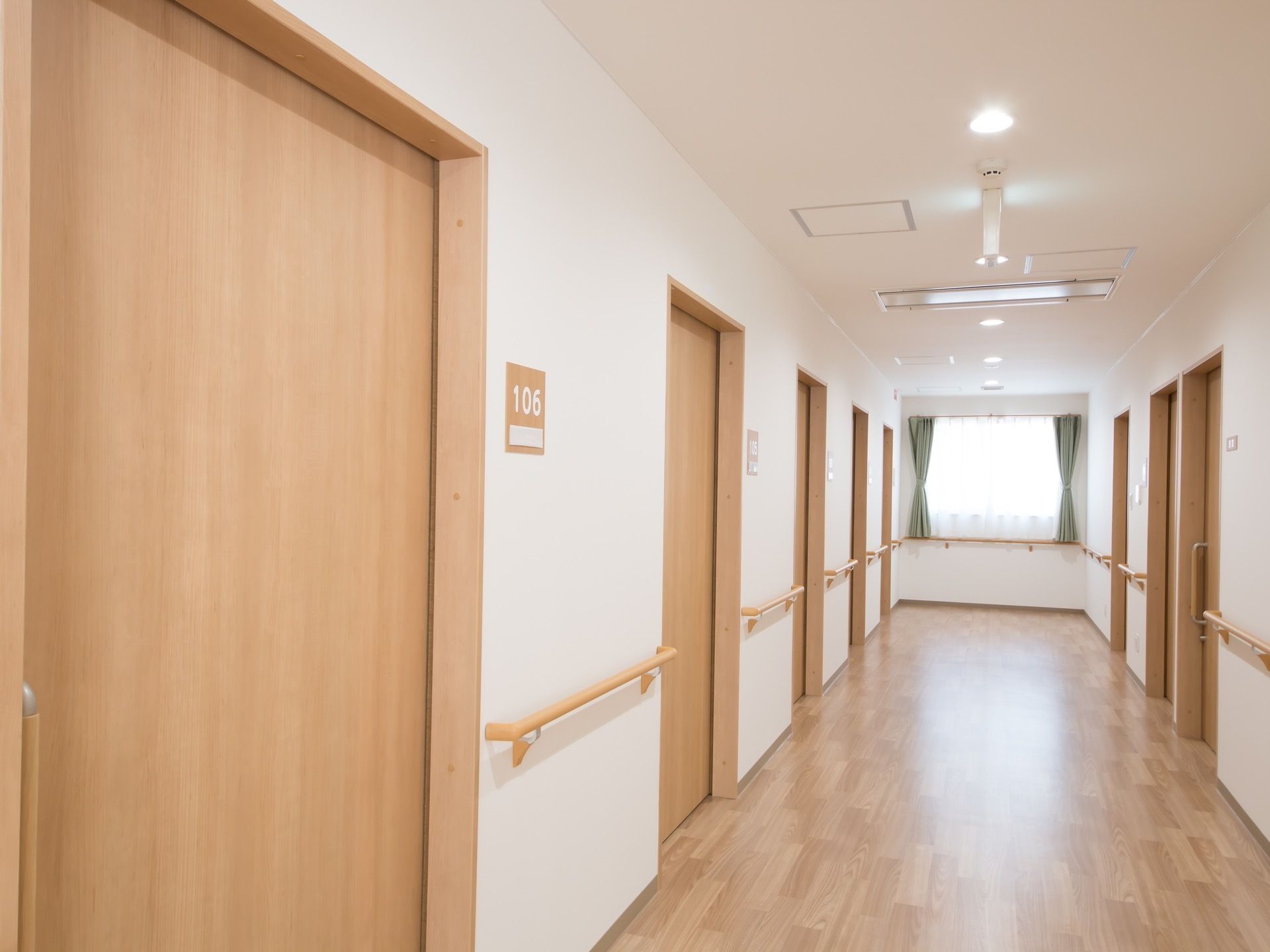 広々とした廊下である。居室が両サイドに並んでいる。壁に沿って手すりが取りつけられている。窓が突き当りに設けられている。