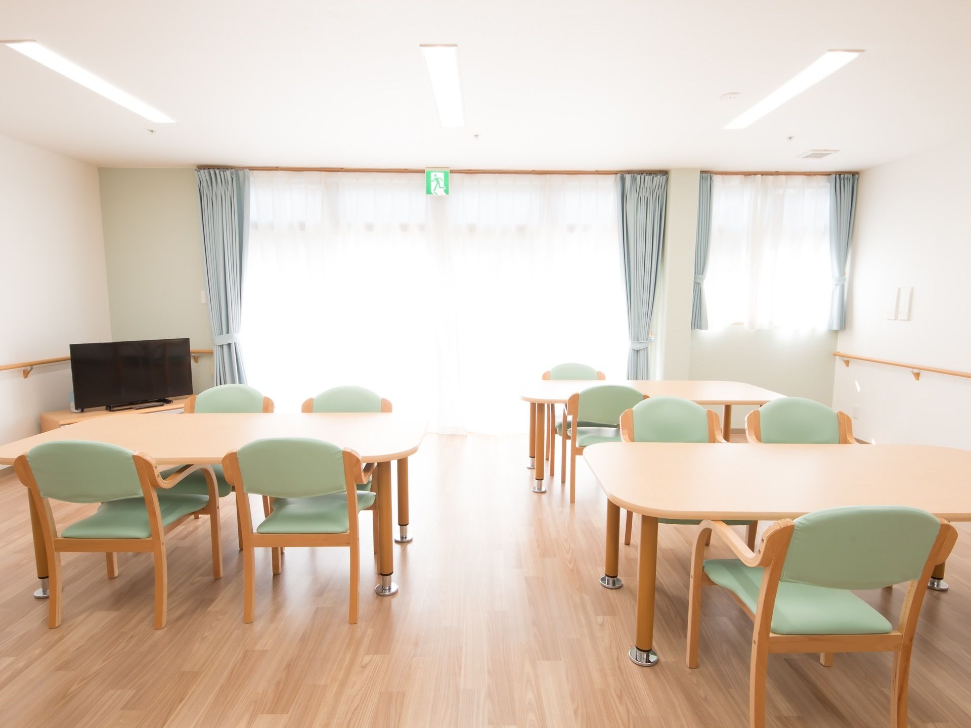 明るい自然光が窓から差し込む広いリビングである。４人掛けテーブルが並べられており、テレビが置かれている。