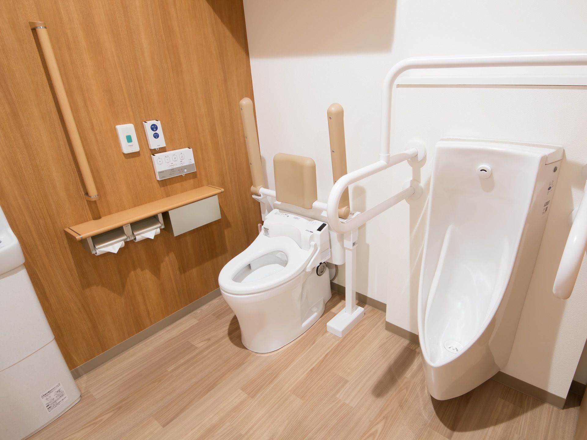 車椅子対応の広いトイレである。背もたれ付きのアームレストや手すりが便器に完備されている。呼び出しボタンや洗面台が設置されている。