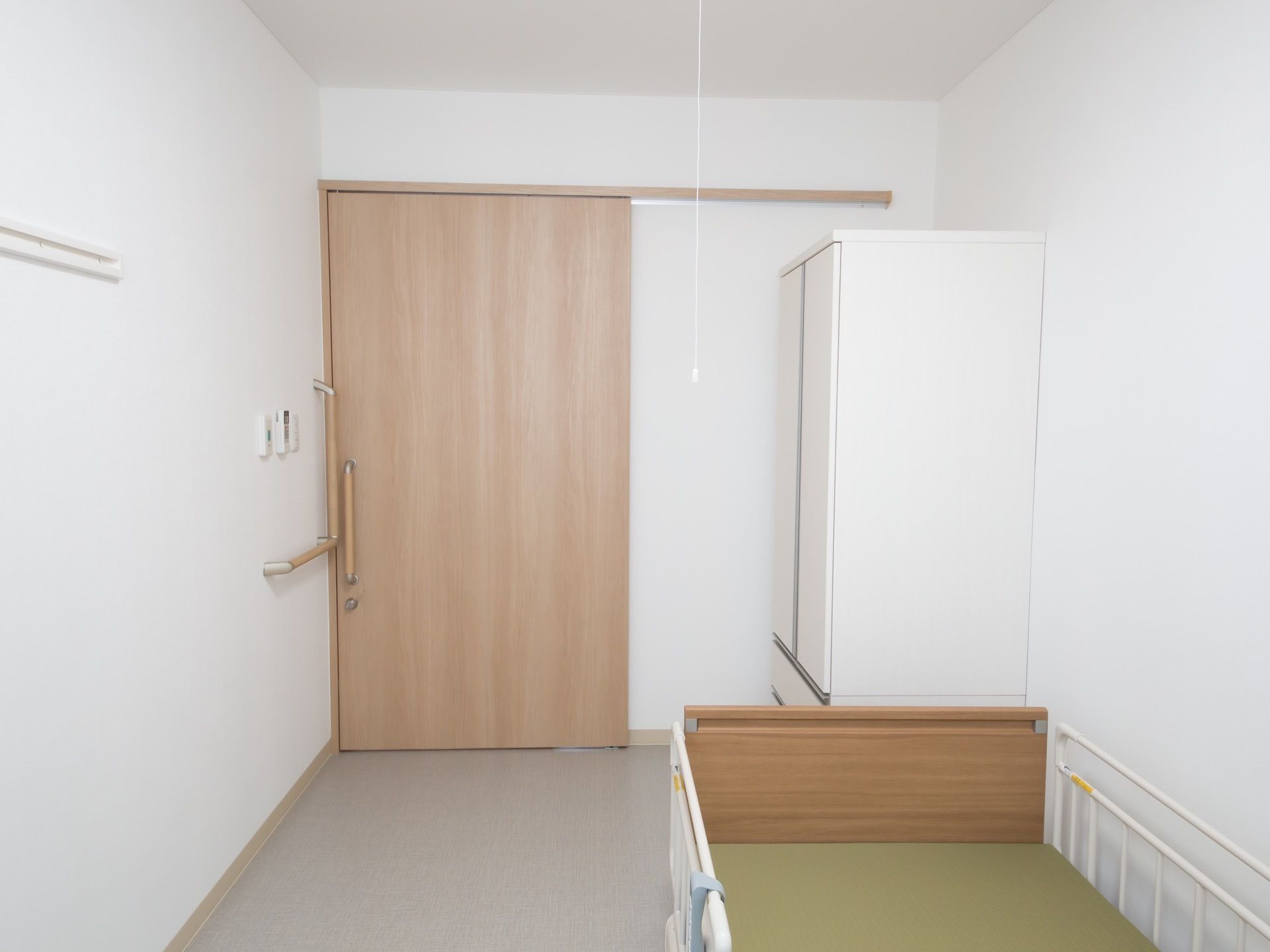収納家具や介護ベッド、照明が完備された個室の居室である。壁には、L型手すりやハンガーフックが設置されている。