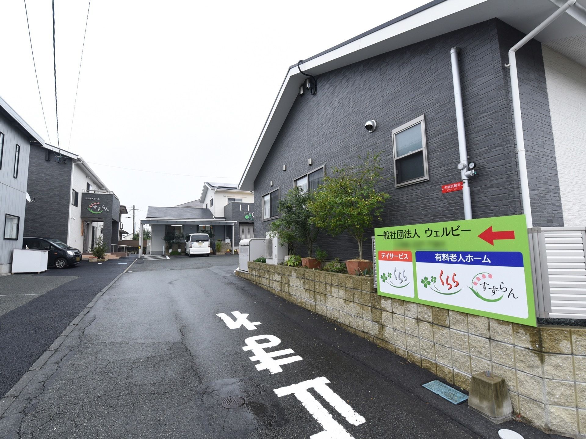 道路から駐車場への入口付近に、案内板が立っており、矢印でホームの方向を示している。デイサービスと有料老人ホームが建ち並んでいる。
