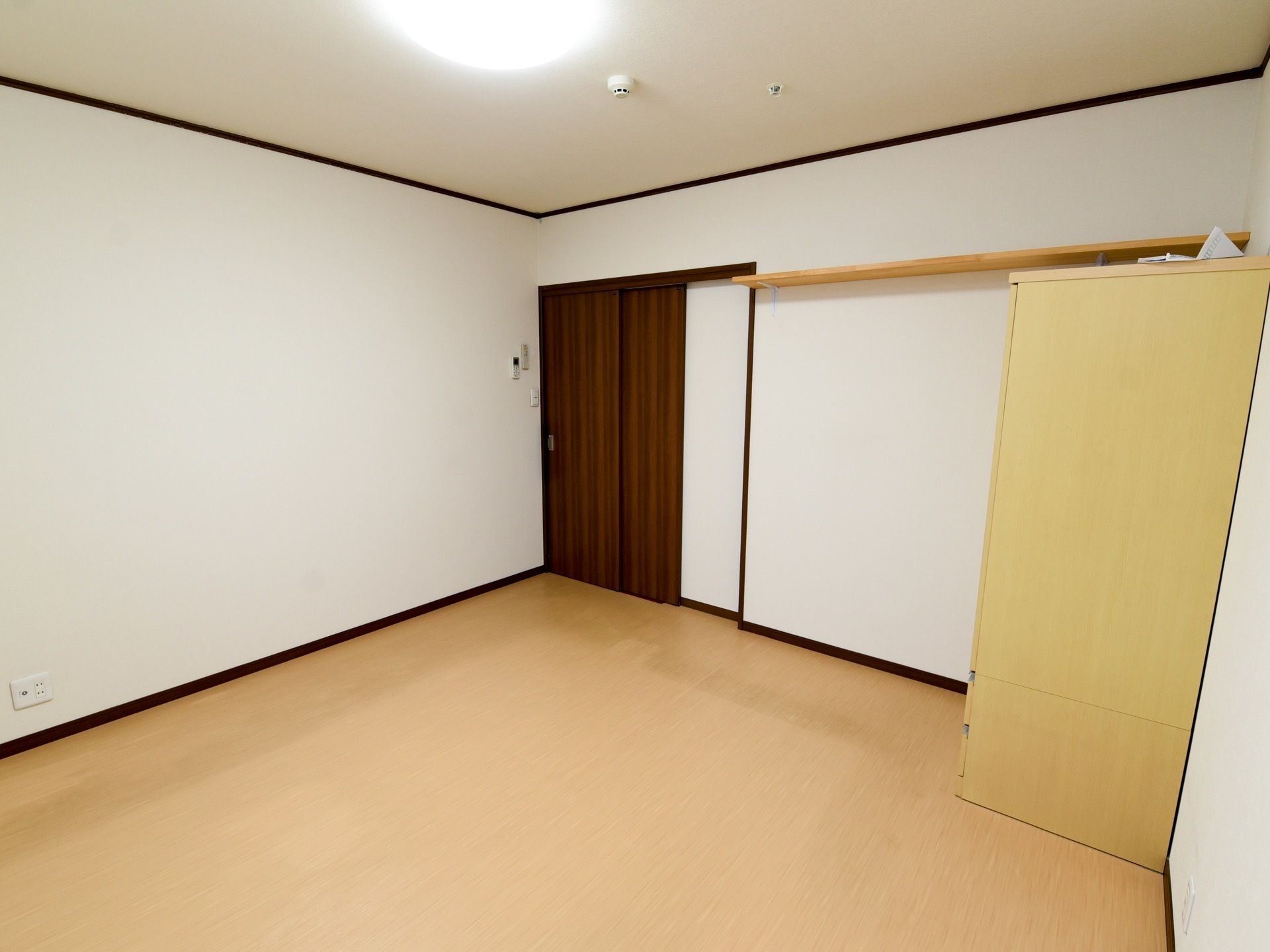 床はベージュ、壁はホワイトで、ダークブラウンの扉がついた居室。入口の近くに、リモコンホルダーが設置してある。