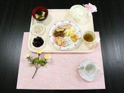 ご飯やみそ汁のほか、数種類のおかずの盛り合わせがお盆の上に並んでいる。ランチョンマットの上に花が添えられている。