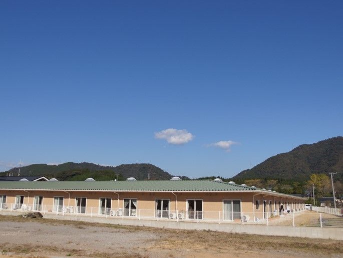 青空の下、緑が映える山々に囲まれた平屋建ての施設建物