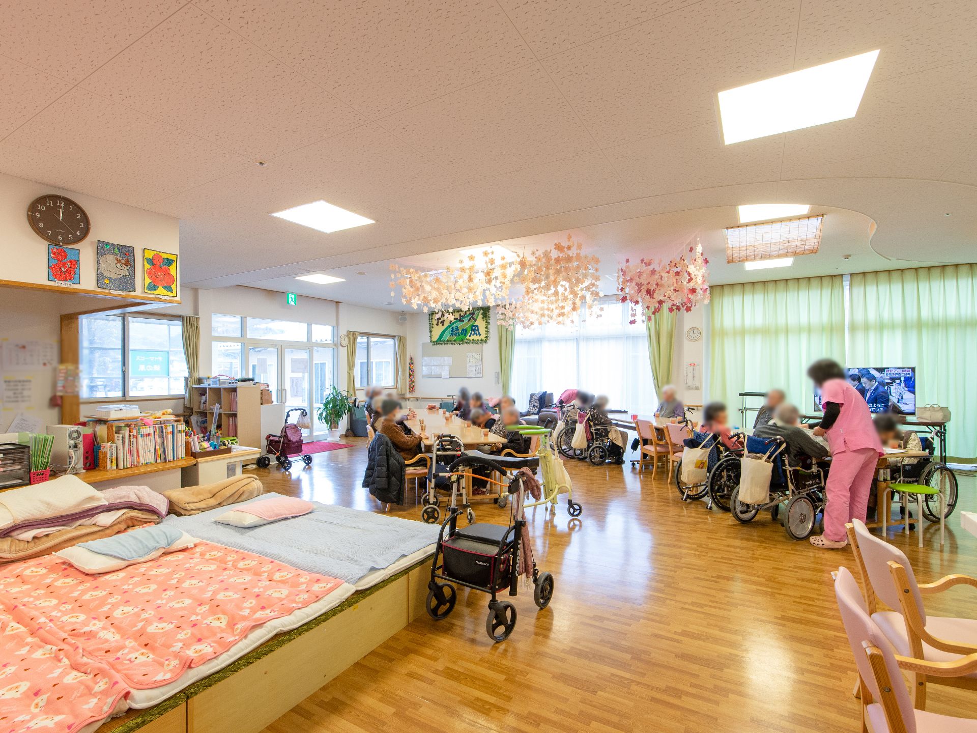 共有スペースに多くの入居者様が集まっている。ピンク色の服を着た女性スタッフが入居者様に声をかけている。