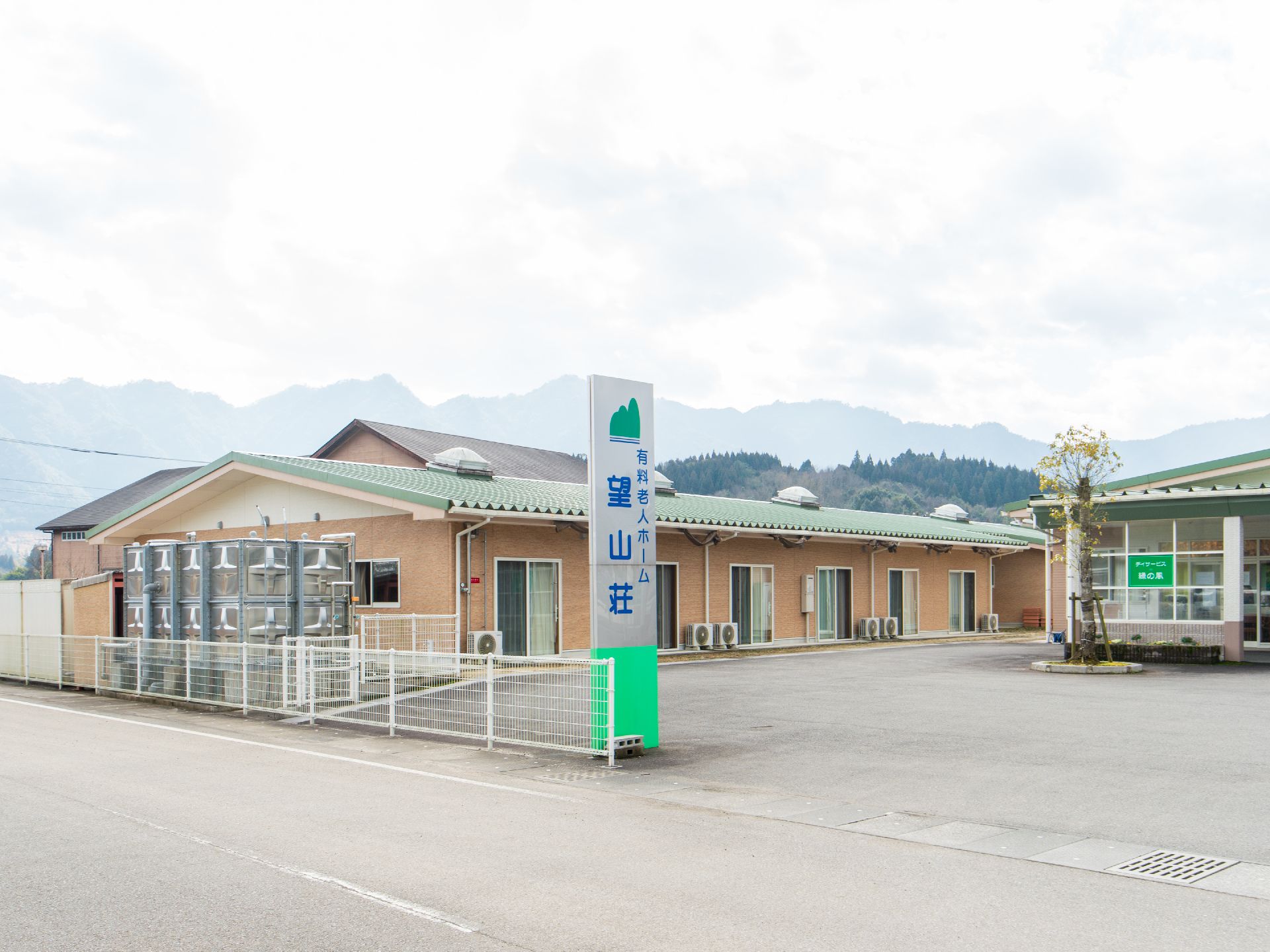 遠近の山並みが見えるのどかな環境に立地している施設である。道路から敷地への入口付近に、施設名称が書かれた看板が設置されている。