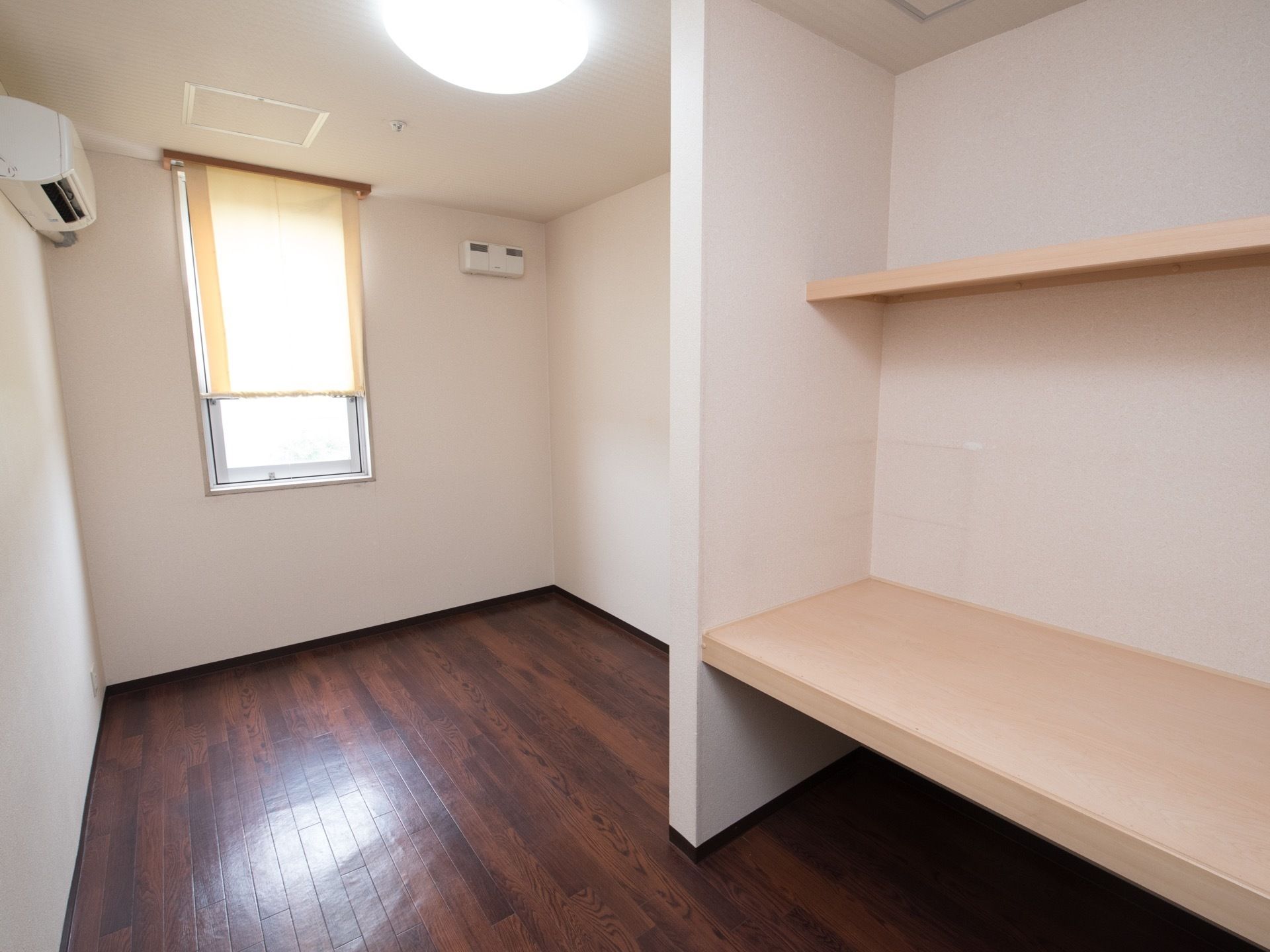 居室の床は落ち着いたダークブラウンのフローリングがである。窓にはクリーム色のロールスクリーンが付けられている。エアコン、収納棚もある。