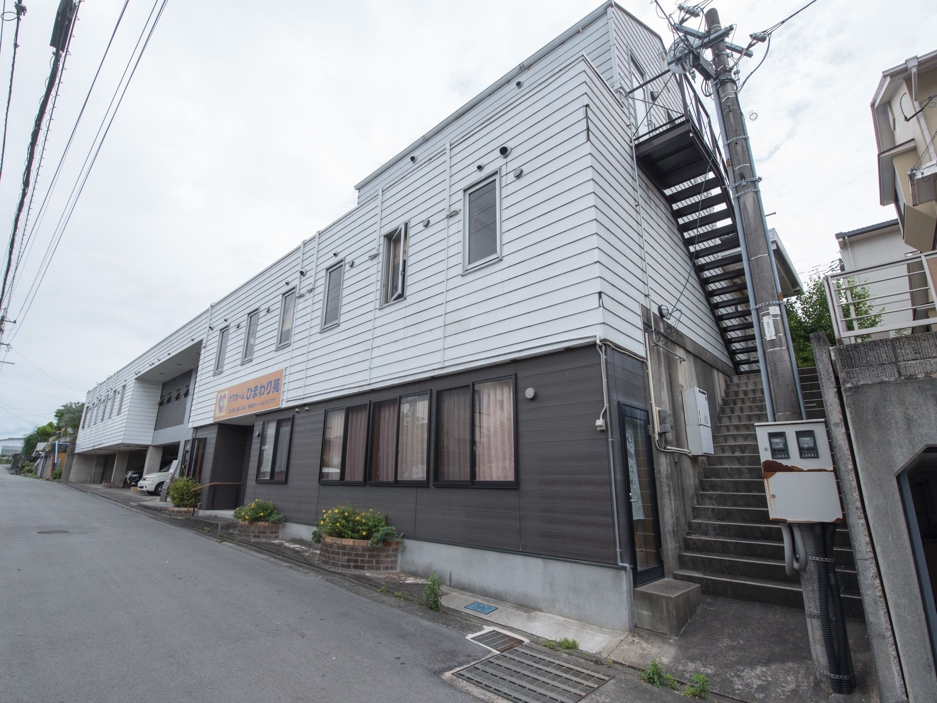 施設は3階建てで、右側には外階段がある。1階の入口付近には手すりが取り付けられている。植え込みもあり、花が咲いている。