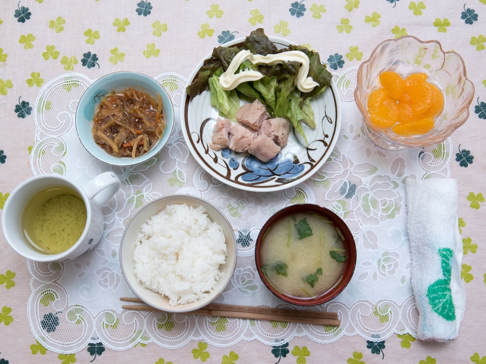 白いレースのランチョンマットの上に食事が配膳されている。この日はご飯、お味噌汁、きんぴらごぼう、お肉、ミカンのメニューである。