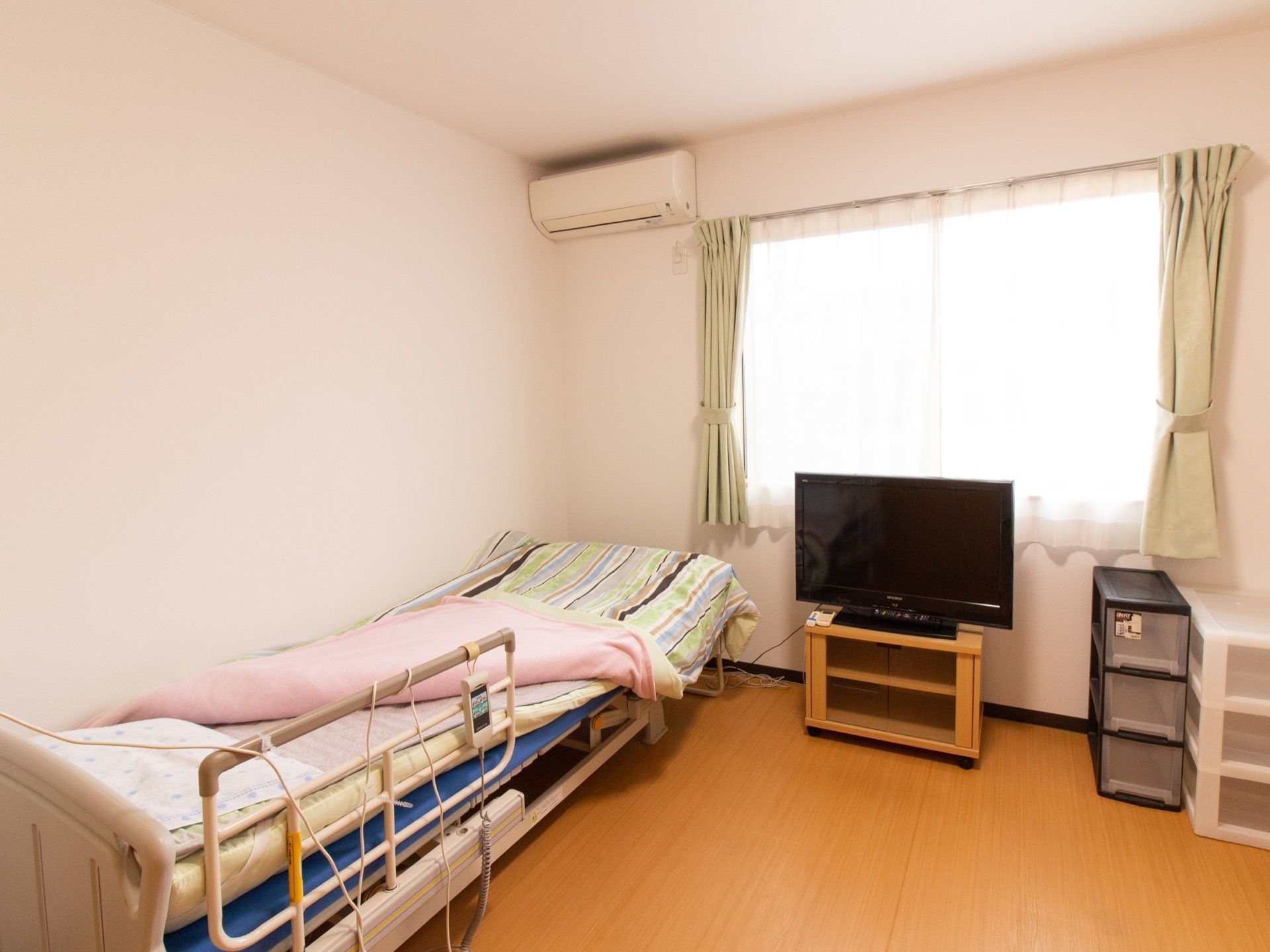 白い壁に窓やエアコンが完備されている居室には、木目調のフラットなフローリングに介護用ベッドが設置されている。