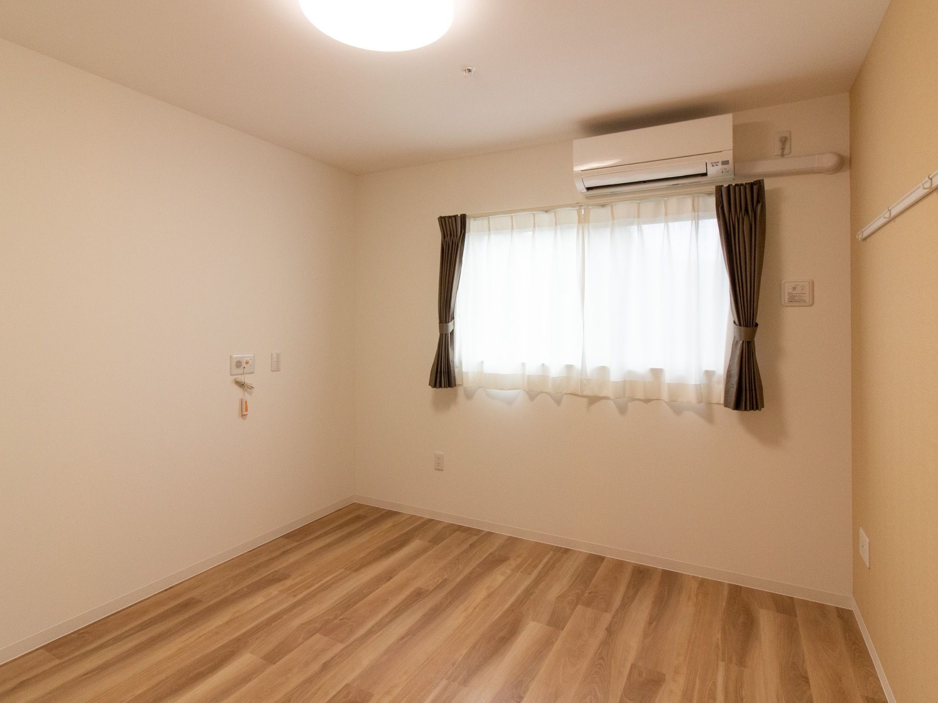 居室は、木目調のフローリングになっている。白い壁にはカーテンが引かれた窓やエアコン、呼び出しブザーが設置されている。