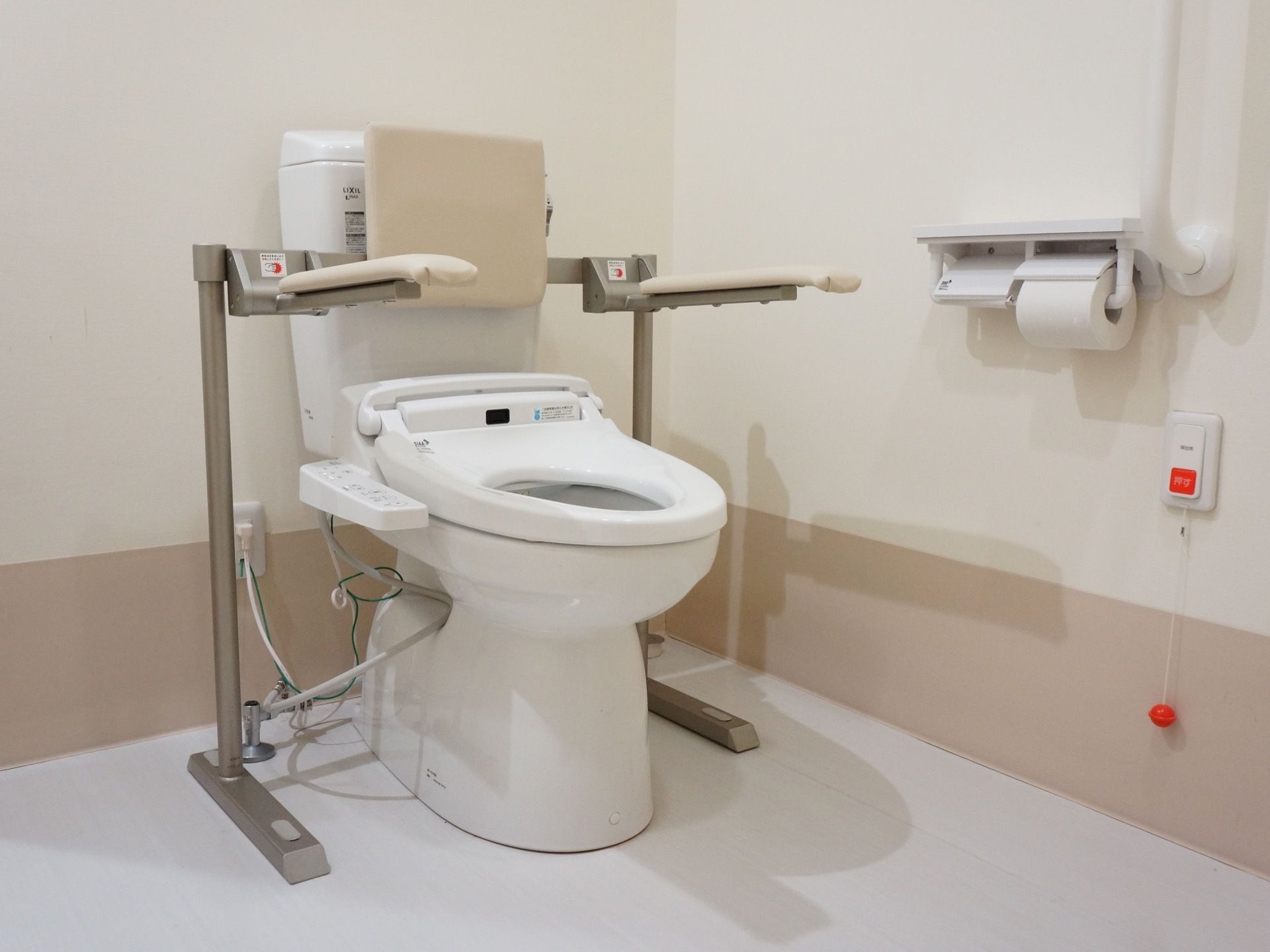 白色を基調としたトイレの個室である。便器には肘掛けや背もたれが備わり、壁には手すりが取り付けられている。