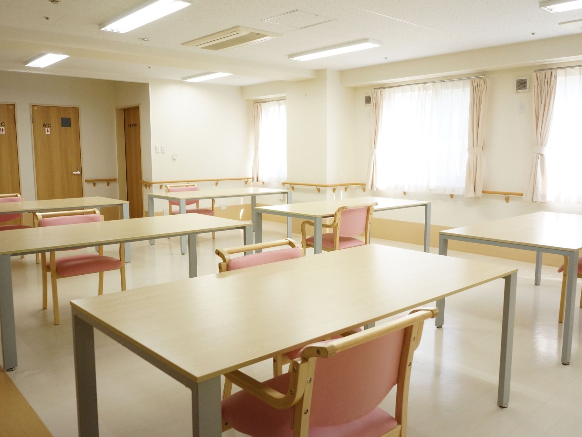 窓がある明るい室内に、長方形のテーブルが並び、背もたれと肘掛けが付いた椅子が備えられている。壁には手すりが備わっている。