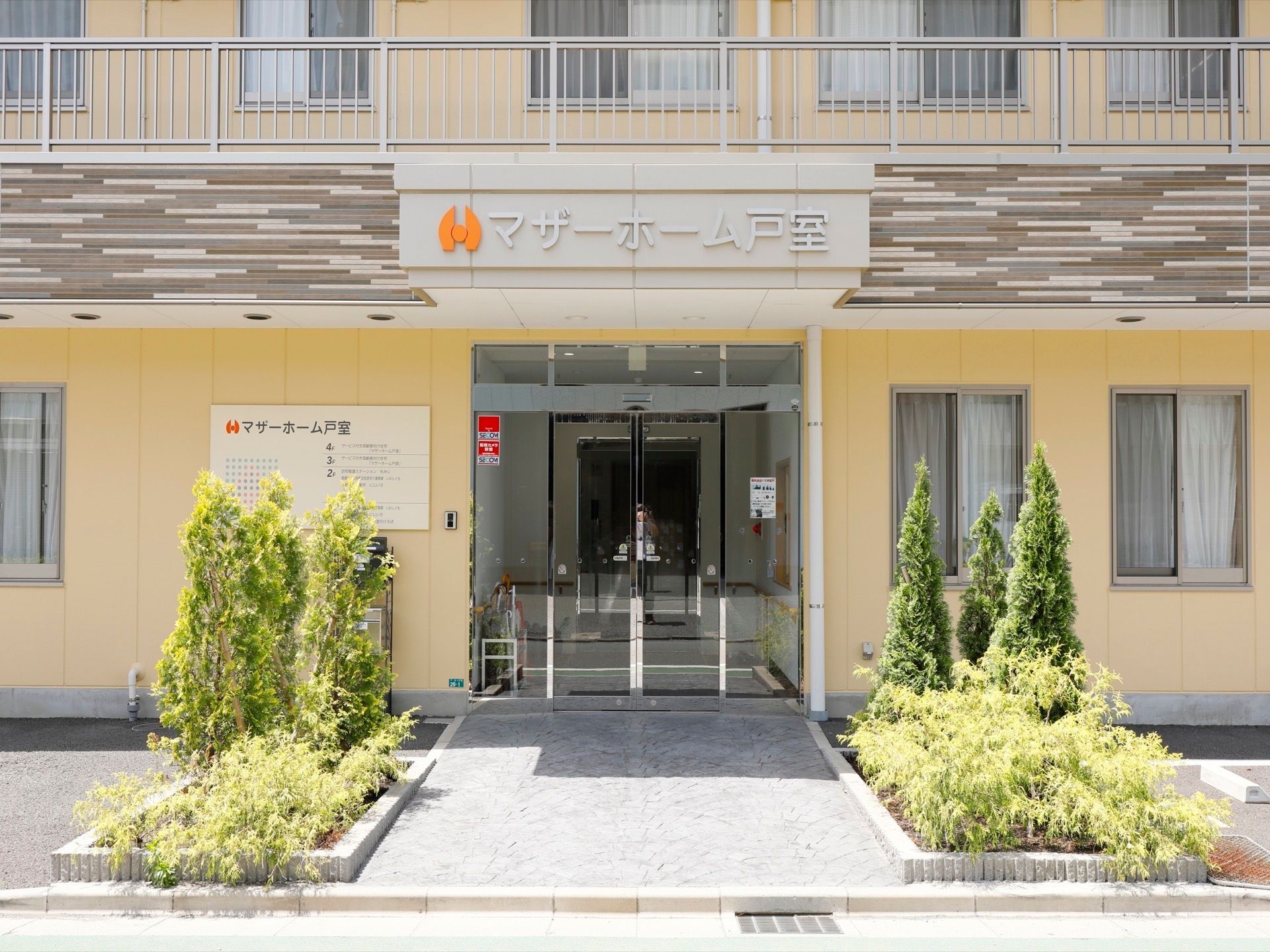 自動ドアの玄関入り口の上部にはひさしがあり、施設の名称が書かれている。入り口手前の左右に、植栽がされている。