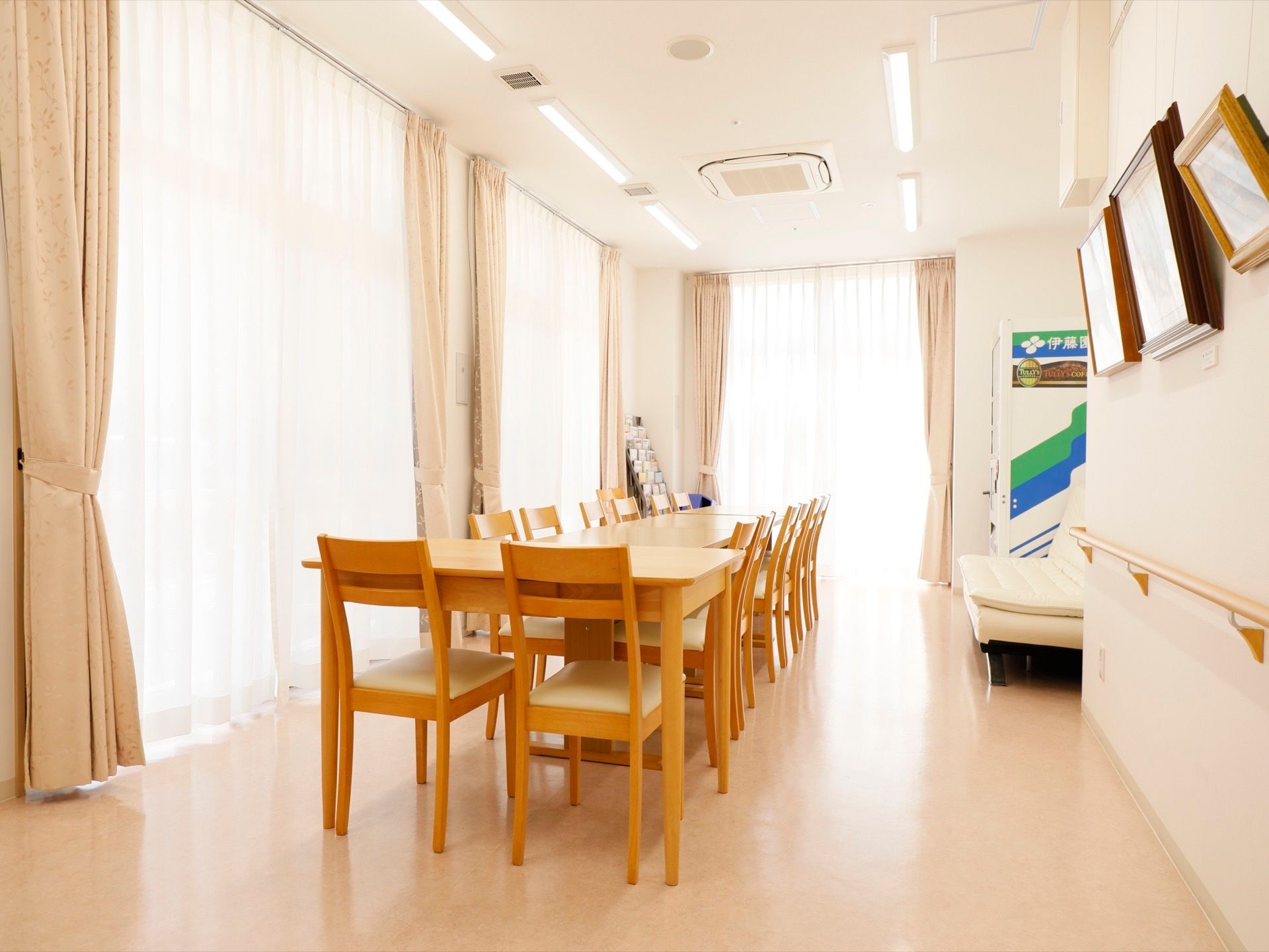 大きな掃き出し窓がいくつもある明るい室内に、テーブルや椅子が並んでいる。自動販売機があり、その横にソファがある。