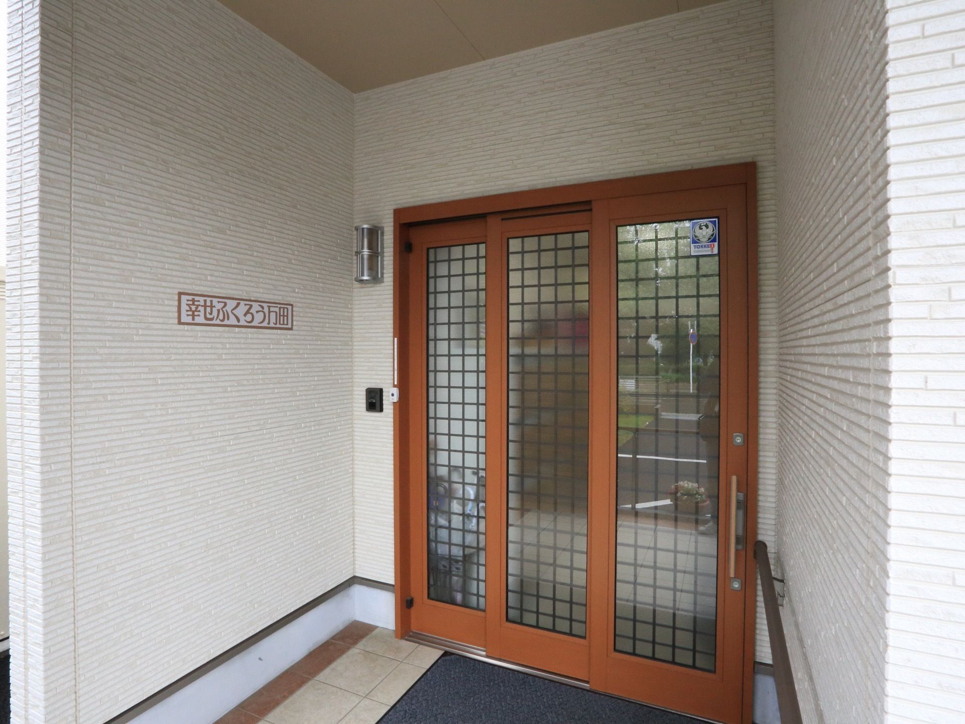 玄関の扉は3枚の連動引き戸を使用しており、開口部が広い。白色のタイル壁に施設名が書かれた看板がある。