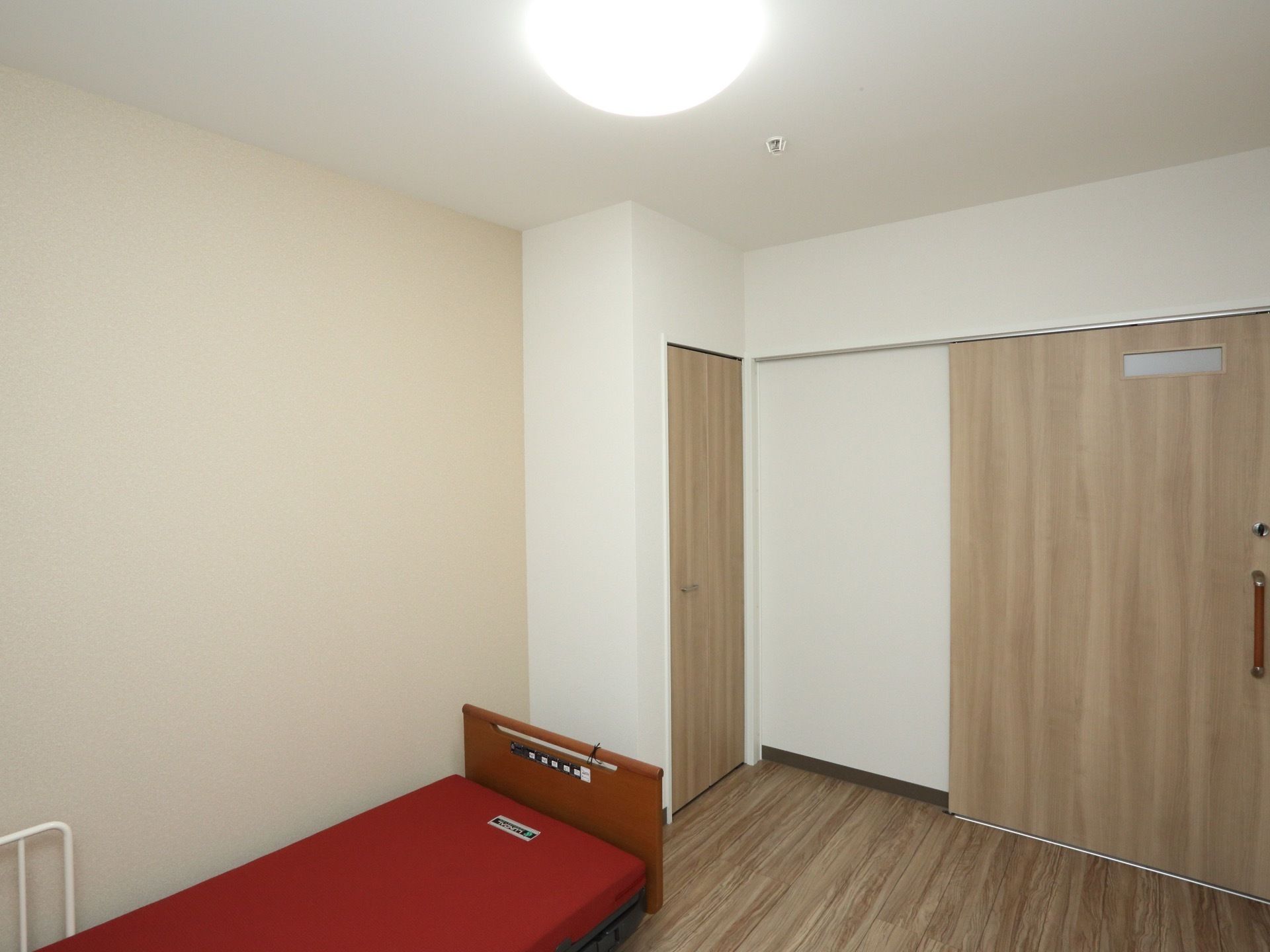 木のぬくもり感あふれる、木目調の床や建具が印象的な居室である。レッド系のベッドが備え付けられている。