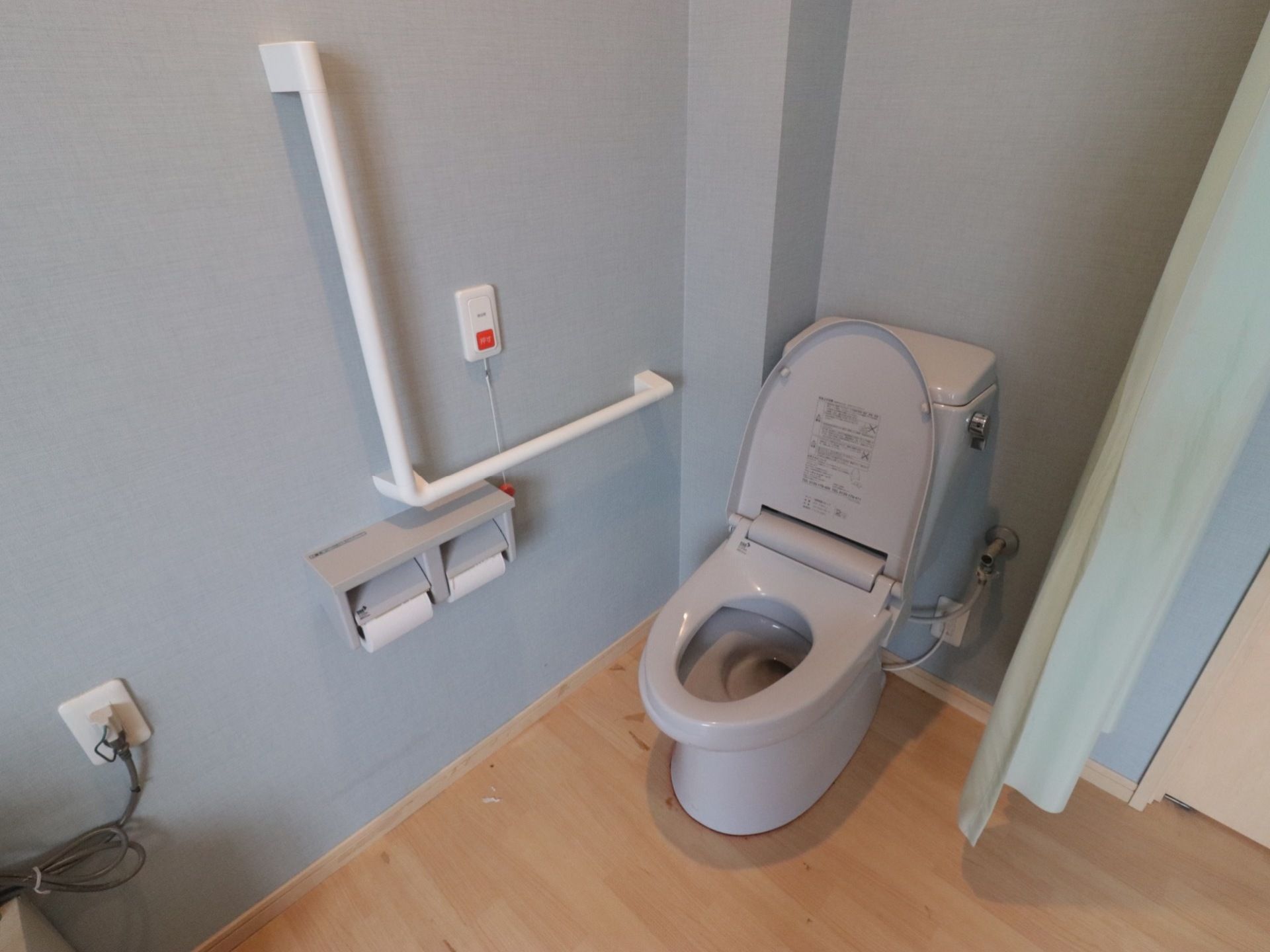 グレー色の洋式便器の便座蓋が開いている。薄いグリーンのカーテンが天井からつるされ、壁に非常ボタンが設置されている。