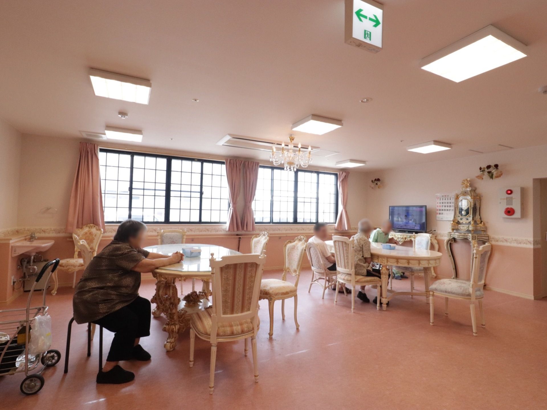 ヨーロピアンスタイルのダイニングテーブルと椅子が置いてある。３名の男女の入居者様が椅子に腰を掛け、テレビを視聴している。