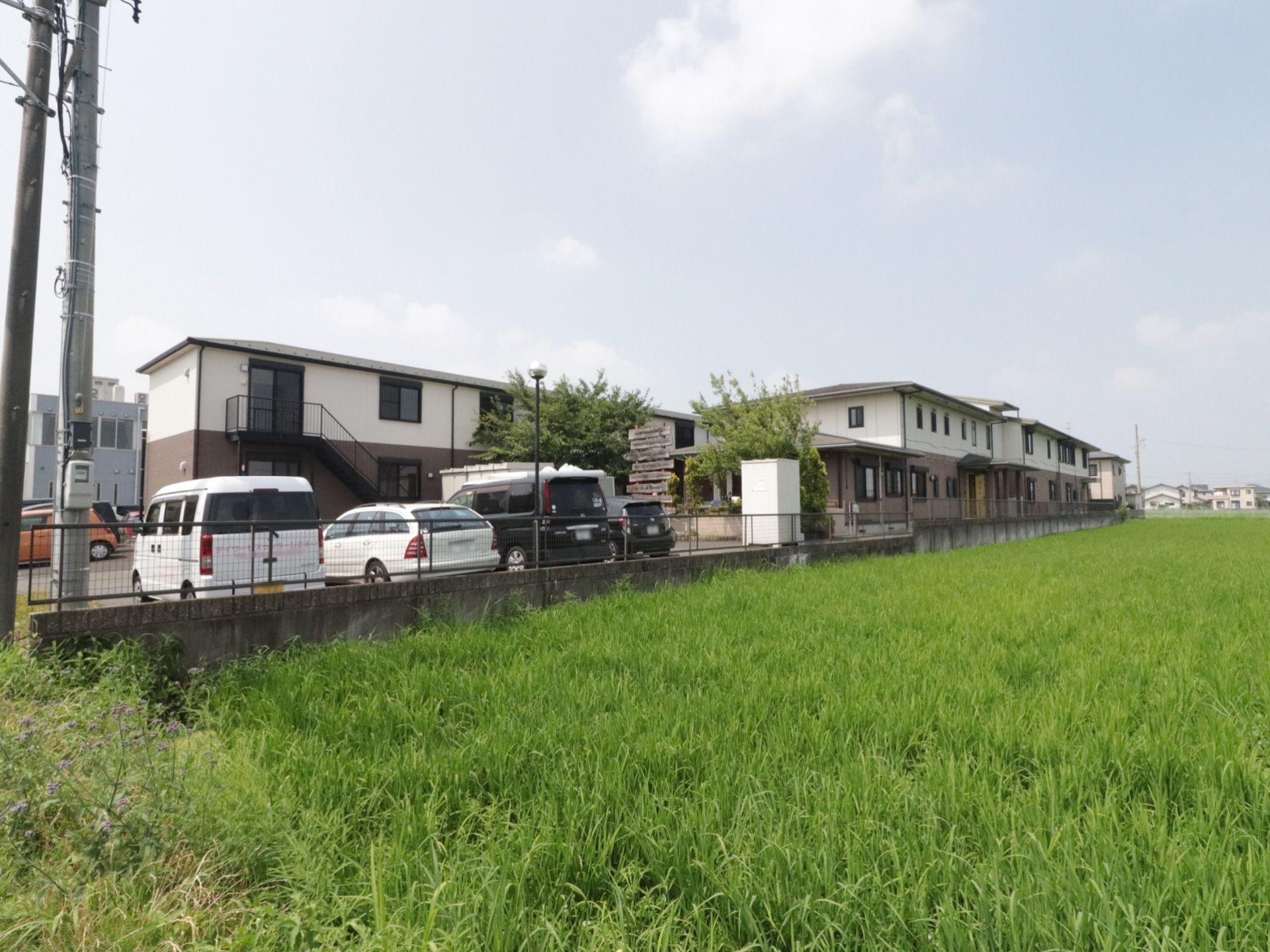 広い田んぼの中に２階建ての建物が建っている。アスファルトで整備された駐車場に数台の車が停められている。