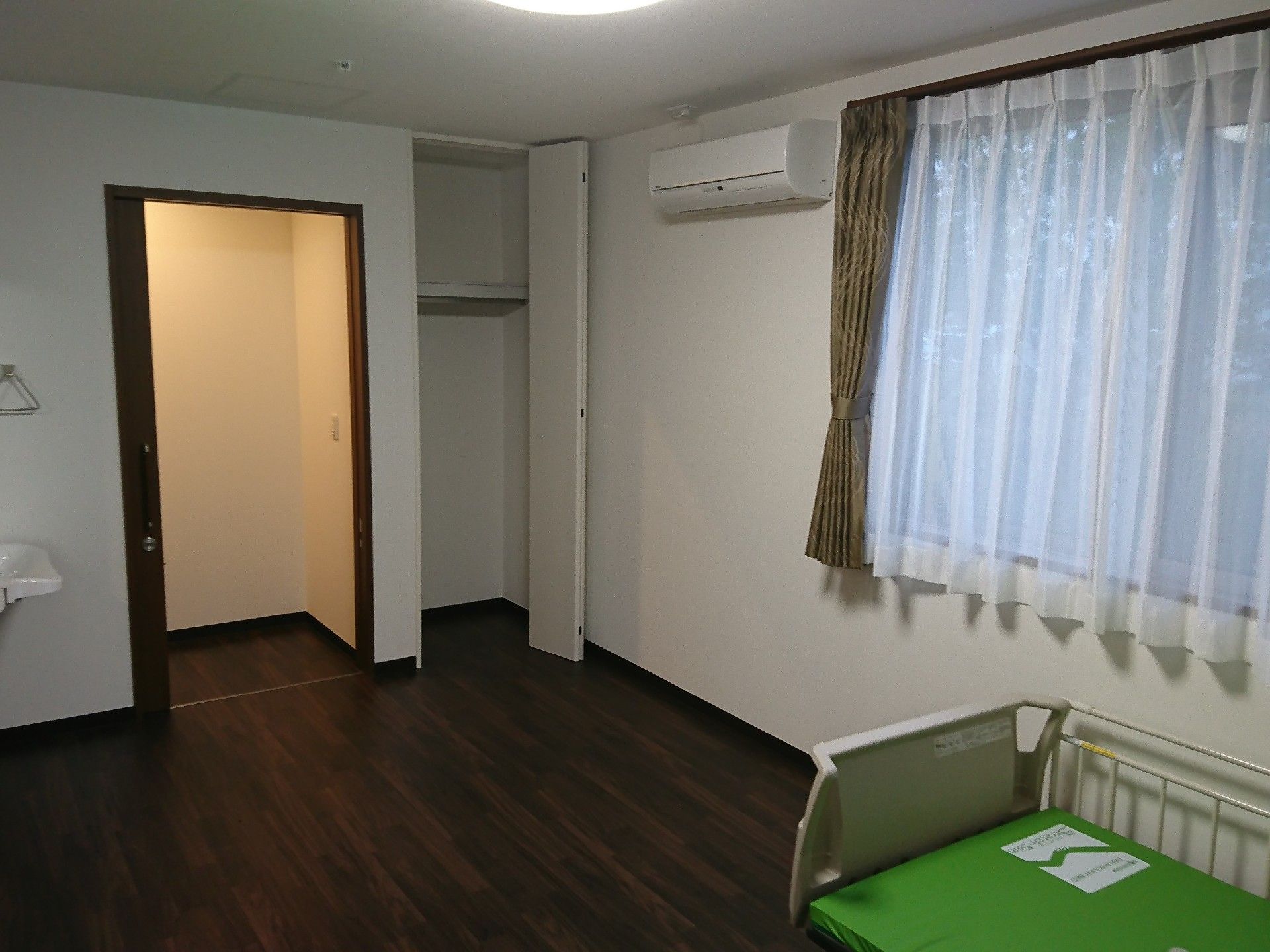落ち着いた色のフローリングの部屋。ベッドが置いてある。窓にはカーテンがついている。手前に洗面台がある。
