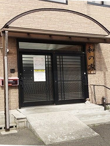 ベージュの外壁の建物の玄関には屋根があり、玄関には手すりやスロープが設置され、玄関のドアには何かが貼られている。