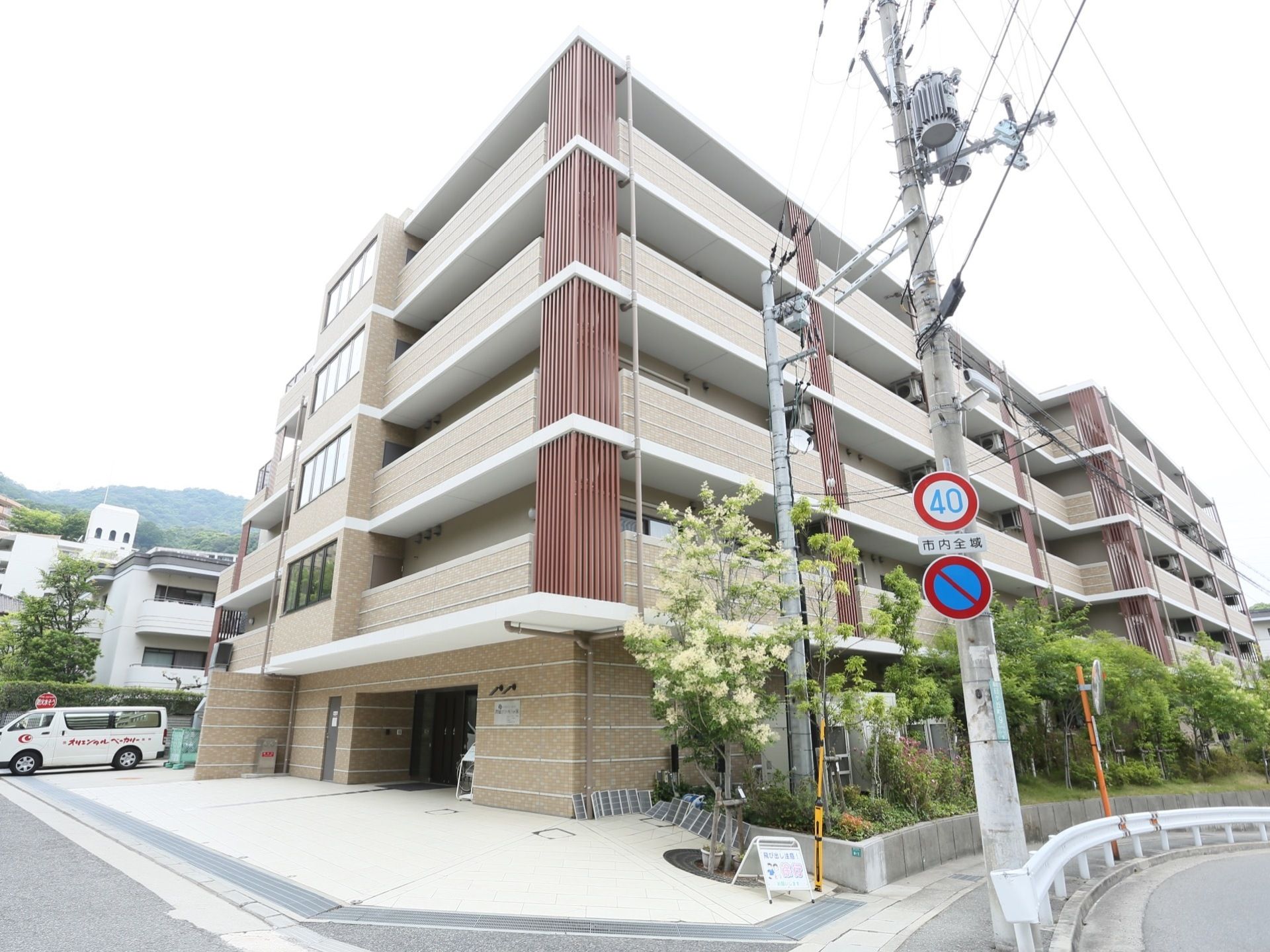 施設はベージュ色の外壁で、５階建てである。建物の横に沿うように花壇が設置されており、側には歩道がある。