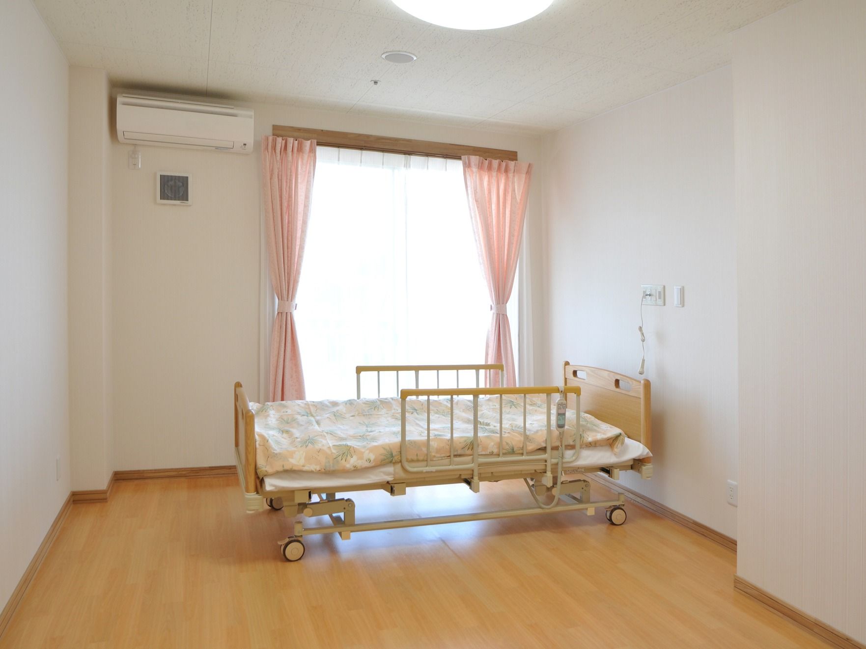 ピンクのカーテンのかかった窓の前にリクライニングベッドが置かれている部屋