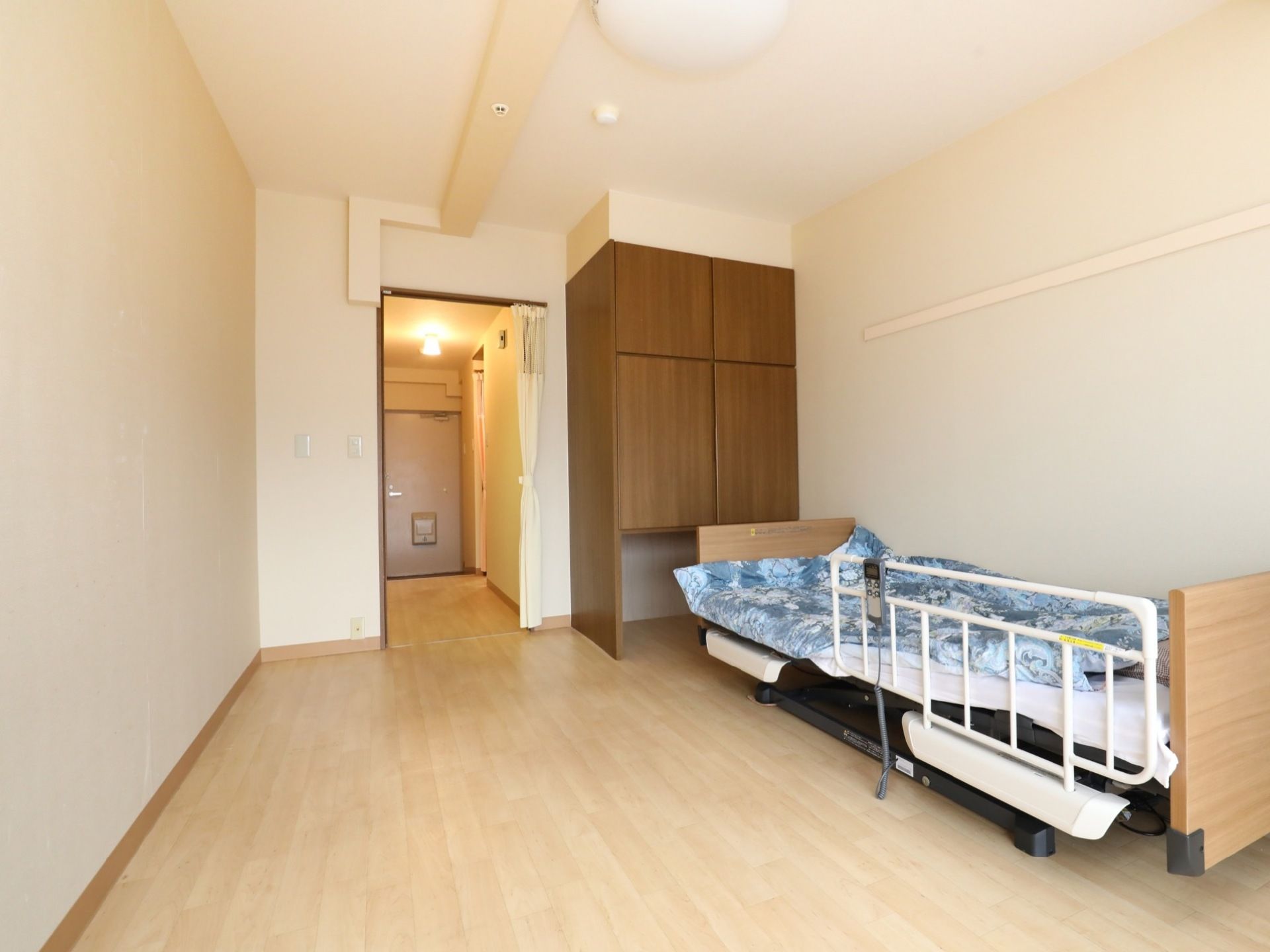 部屋の右側に電動ベッドが置いてあり、その近くには備えつけのクローゼットがある。玄関周辺は、暖色系の照明がついている。