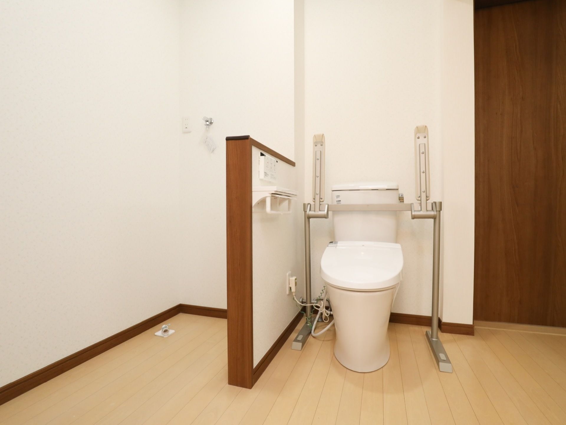 温水洗浄便座を採用したトイレがあり、操作ボタンが壁にある。便器の両側に可動式のひじ掛けを取りつけている。