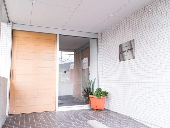 茶色の引き戸の玄関。季節の花々が入居者様をお出迎えする。右手に本施設の銀色のプレートを取り付けている。
