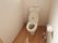 白い壁の明るく清潔感のある洋式トイレである。ブラウン色の木目が使われた床になっており、落ち着いた雰囲気である。